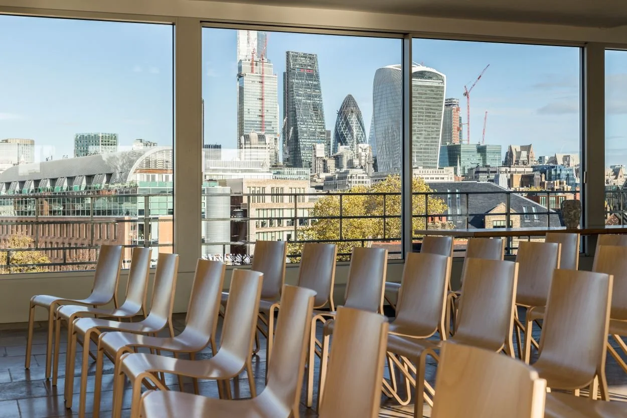 Interior of a conference room or auditorium with multiple wooden chairs arranged facing a large window. The window offers a view of the London skyline, including the Shard, the Walkie Talkie building, and other skyscrapers.