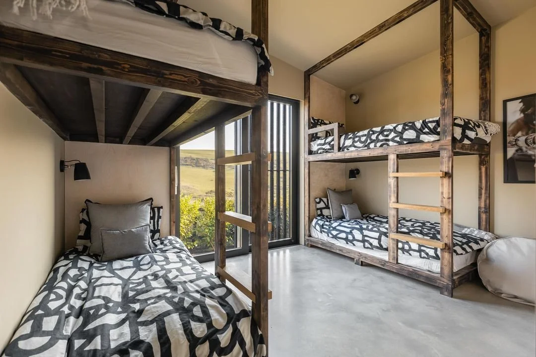 Room with three beds featuring black and white patterned bedding, two on the floor and one on a raised wooden bunk bed, with a large window showing outdoor landscape.
