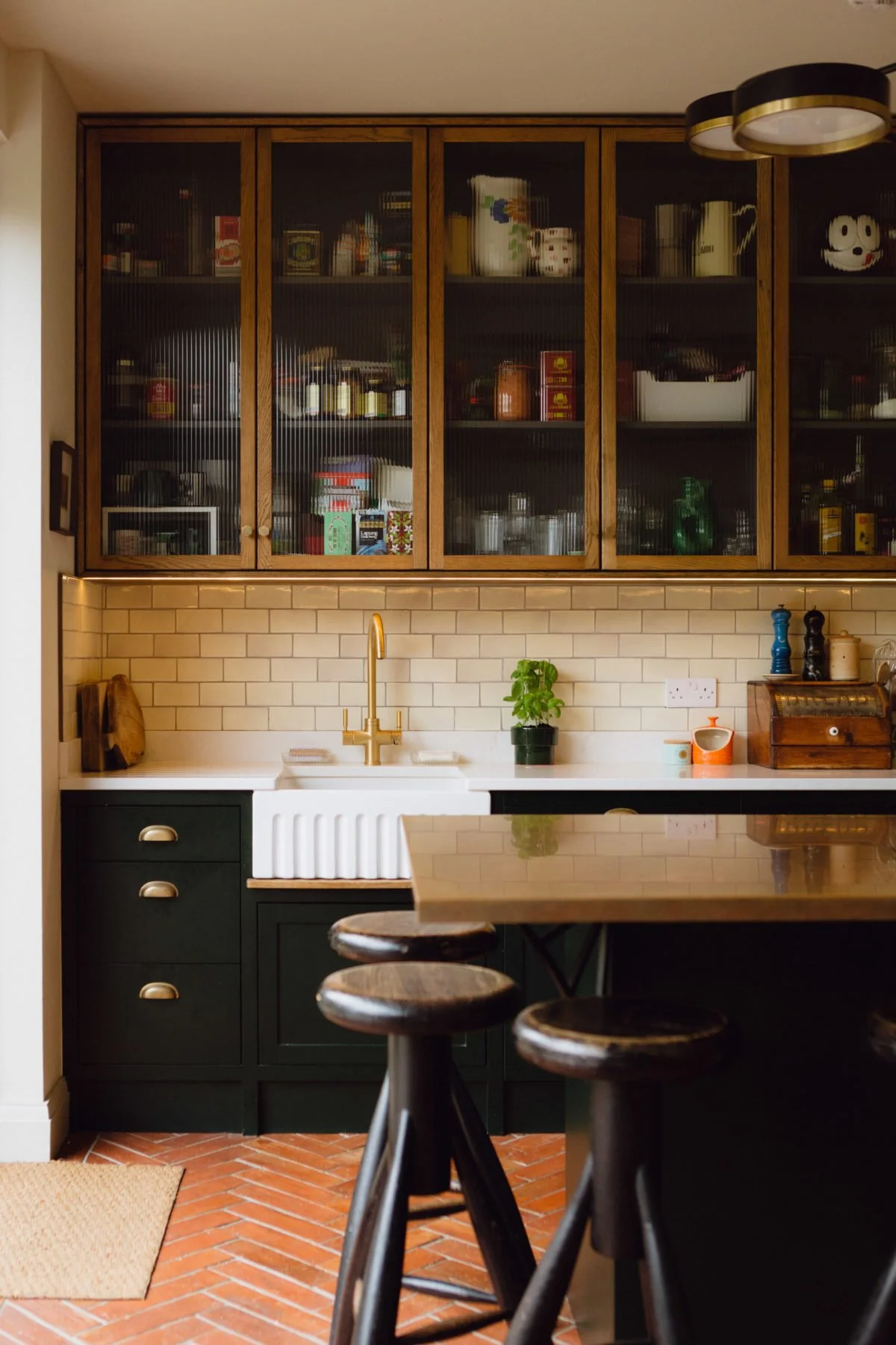 A modern kitchen with dark green lower cabinets, a white farmhouse sink with a gold faucet, and beige subway tile backsplash. Glass-front upper cabinets display dishes and food items. A small potted plant is on the countertop. There is a wooden table