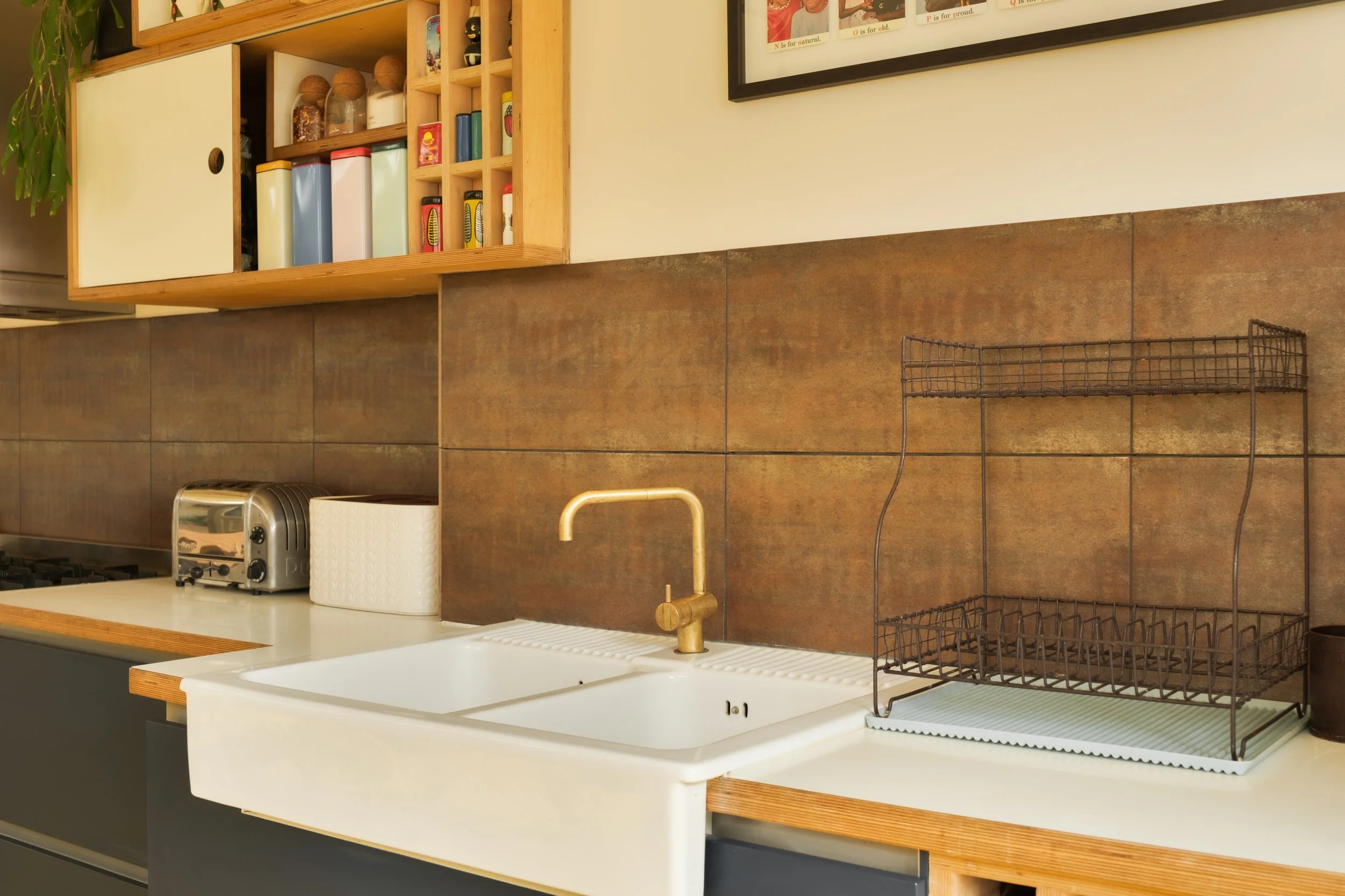 Kitchen countertop with a white farmhouse sink, brass faucet, brown wire dish rack, toaster, and paper towel holder. Wooden shelves with jars and containers above, and a framed picture on the wall.
