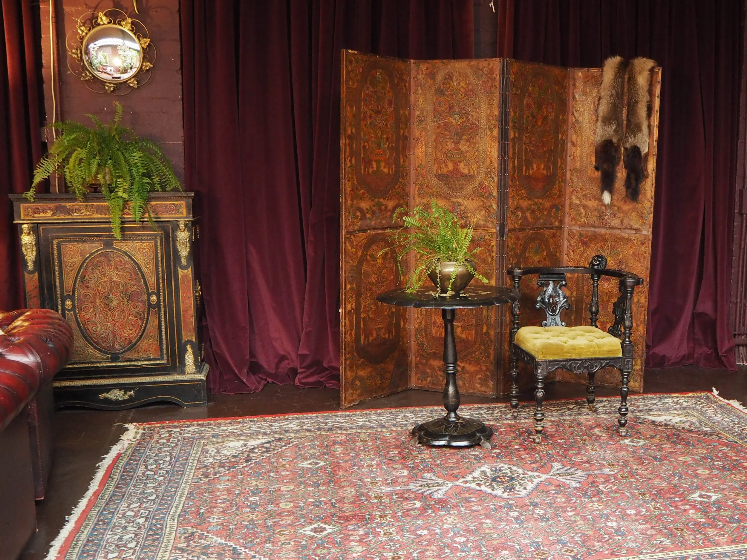 Vintage room with ornate furniture, a patterned rug, burgundy curtains, plants, and animal pelts hanging on a folding screen.