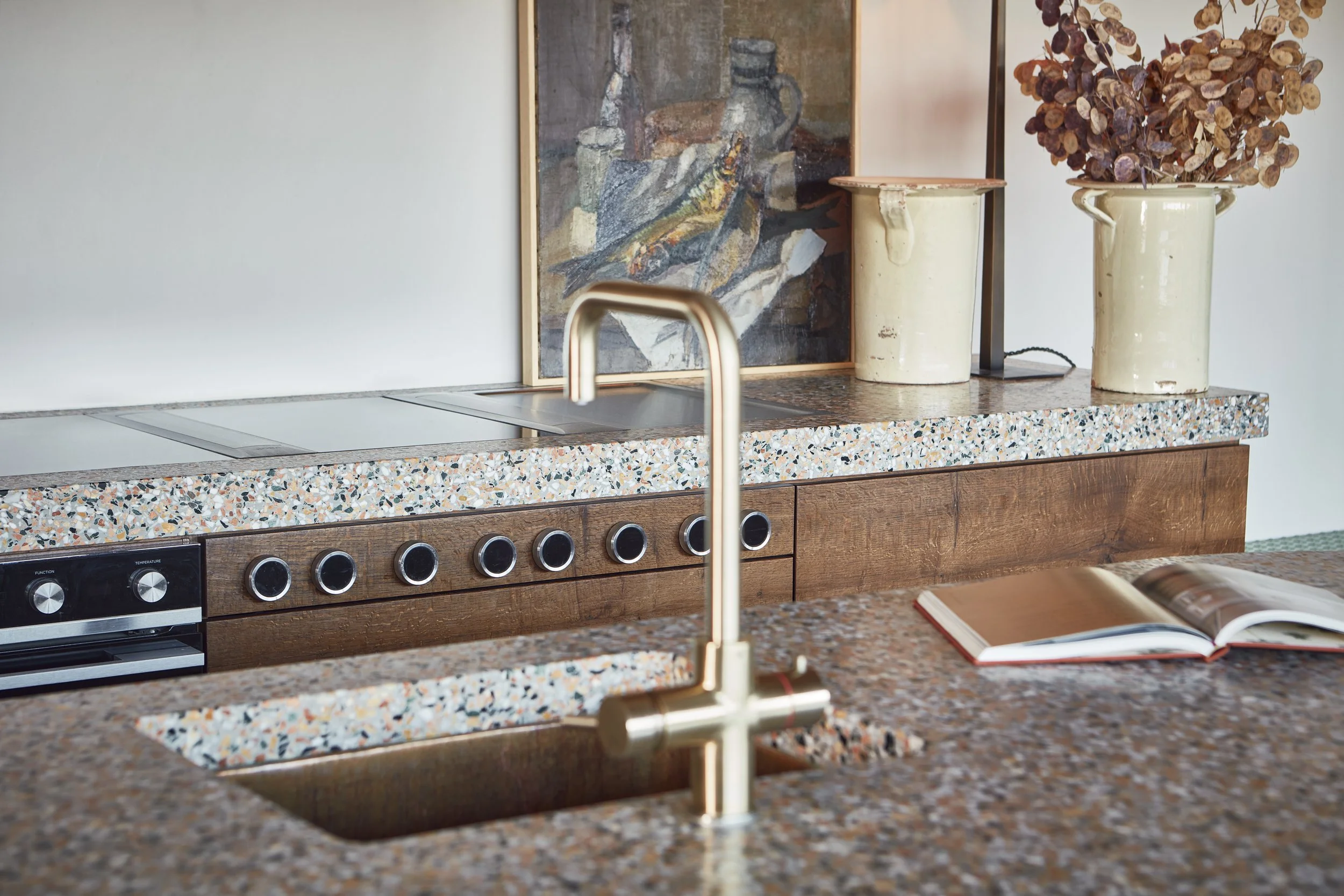 A kitchen countertop with a stainless steel sink, a modern brass faucet, and a colorful terrazzo surface. Behind the counter, there's a wooden cabinet with a book open on the surface. Two large cream-colored planters, one with dried hydrangeas and th