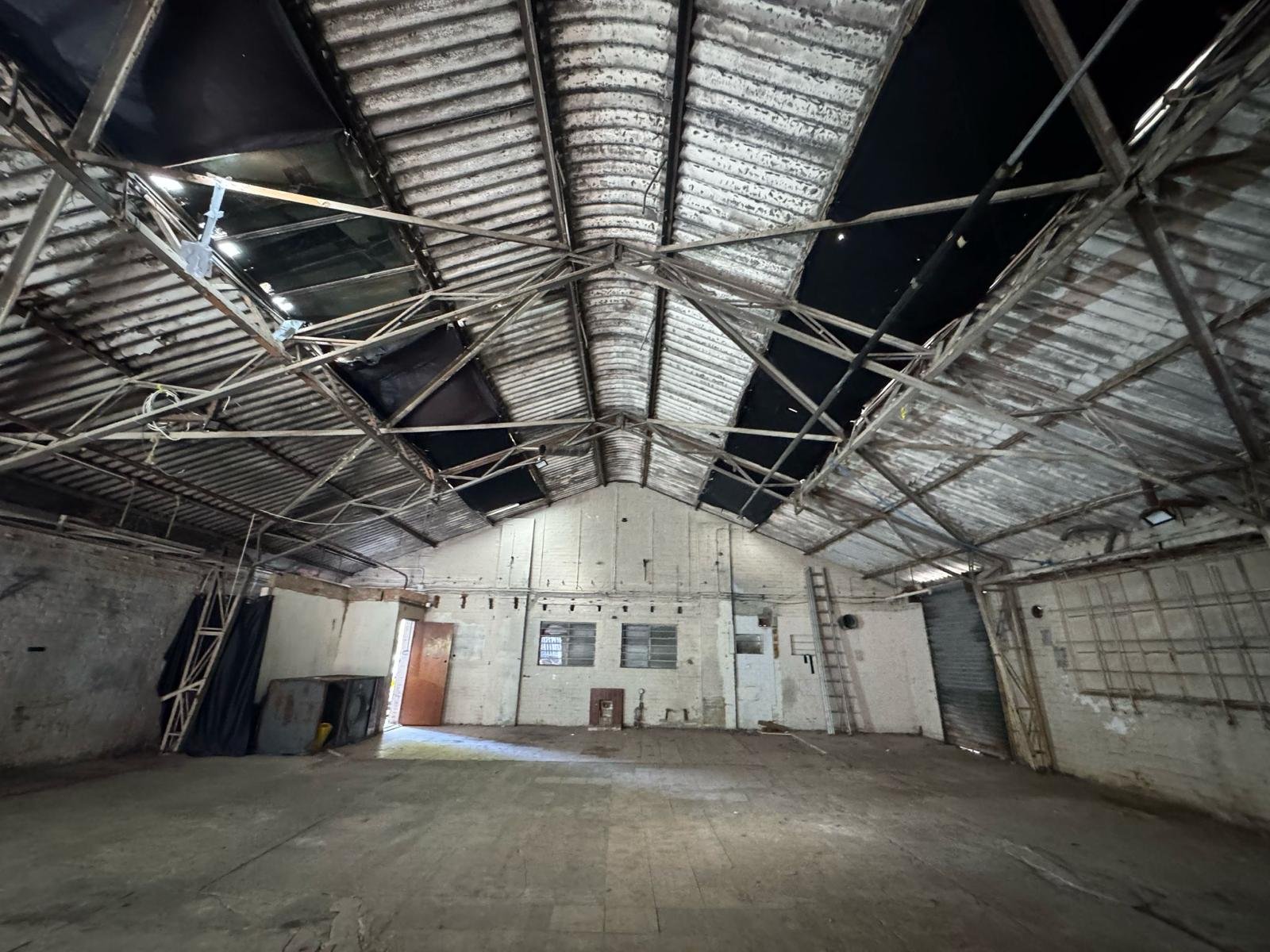 Interior of a large, empty industrial warehouse with high, arched metal roof and exposed metal framing. Ceiling panels are weathered, with some sections missing or damaged. The walls are made of brick and concrete, with small windows and a few doors,