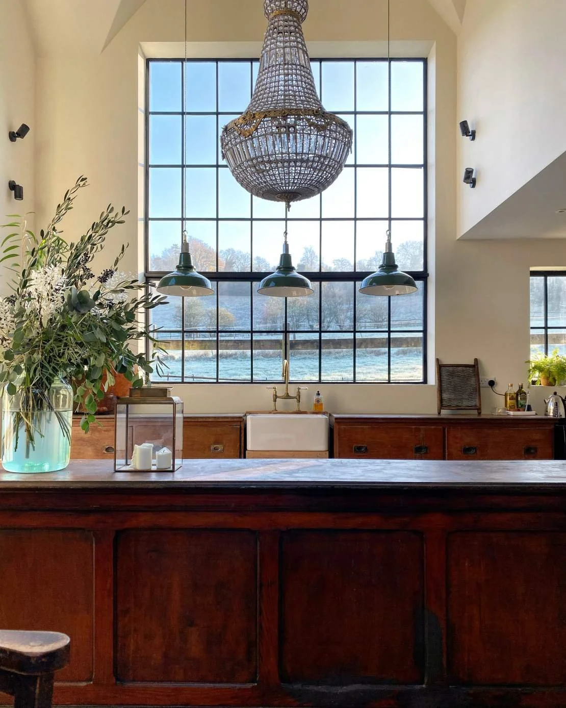 A large, ornate crystal chandelier hanging above a kitchen island with a farmhouse sink, set in front of a large grid-style window. The kitchen features dark wood cabinets, a potted plant, candles, and various bottles on the counter.