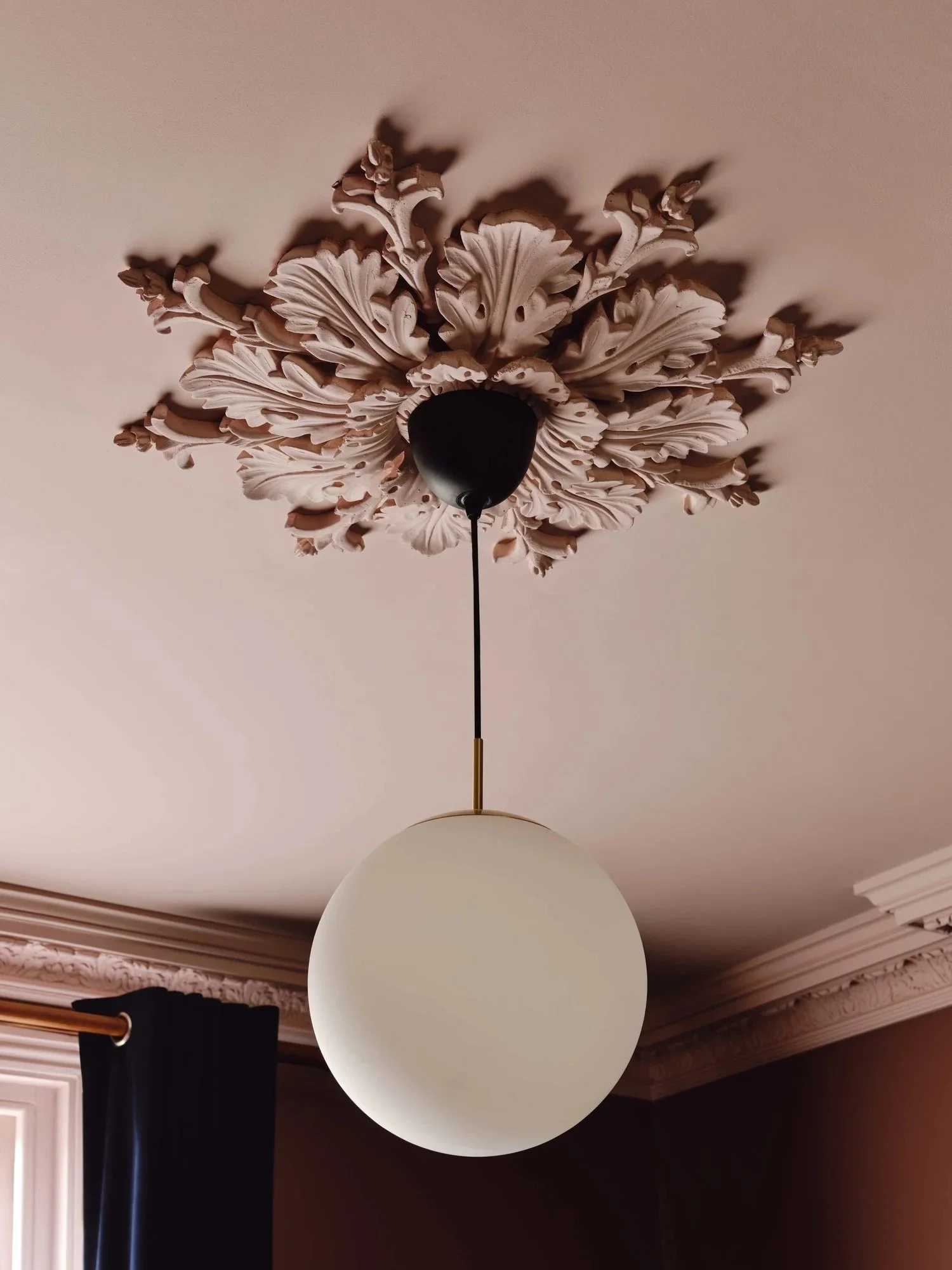 A ceiling light fixture with a frosted glass globe hanging from a black and gold mount, installed on a ceiling with ornate floral and leaf-designed ceiling medallion.