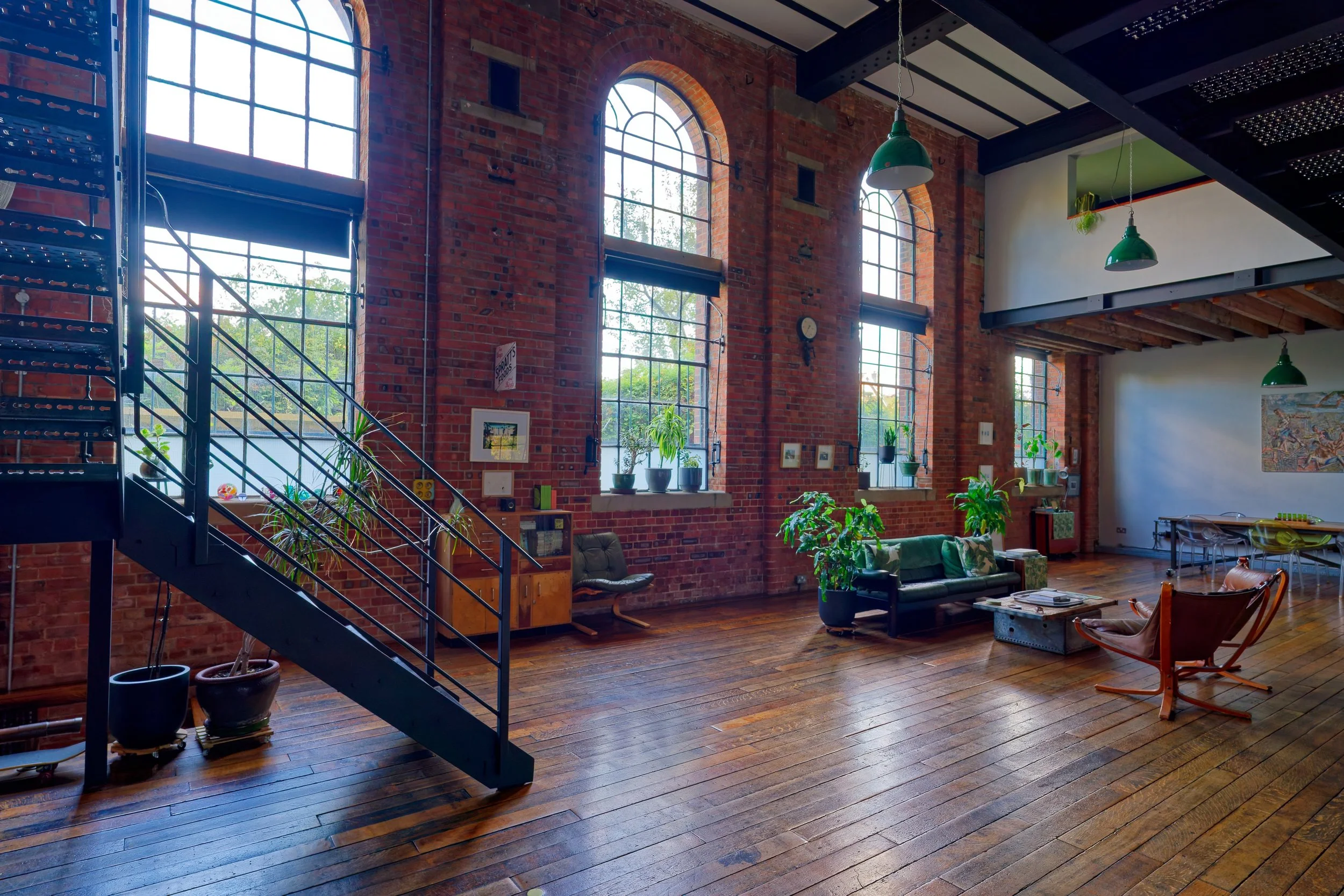 Interior of a spacious loft with large arched windows, exposed brick walls, wooden floors, and green hanging pendant lights. There are plants on the windowsills and furniture including a green sofa and a wooden armchair.