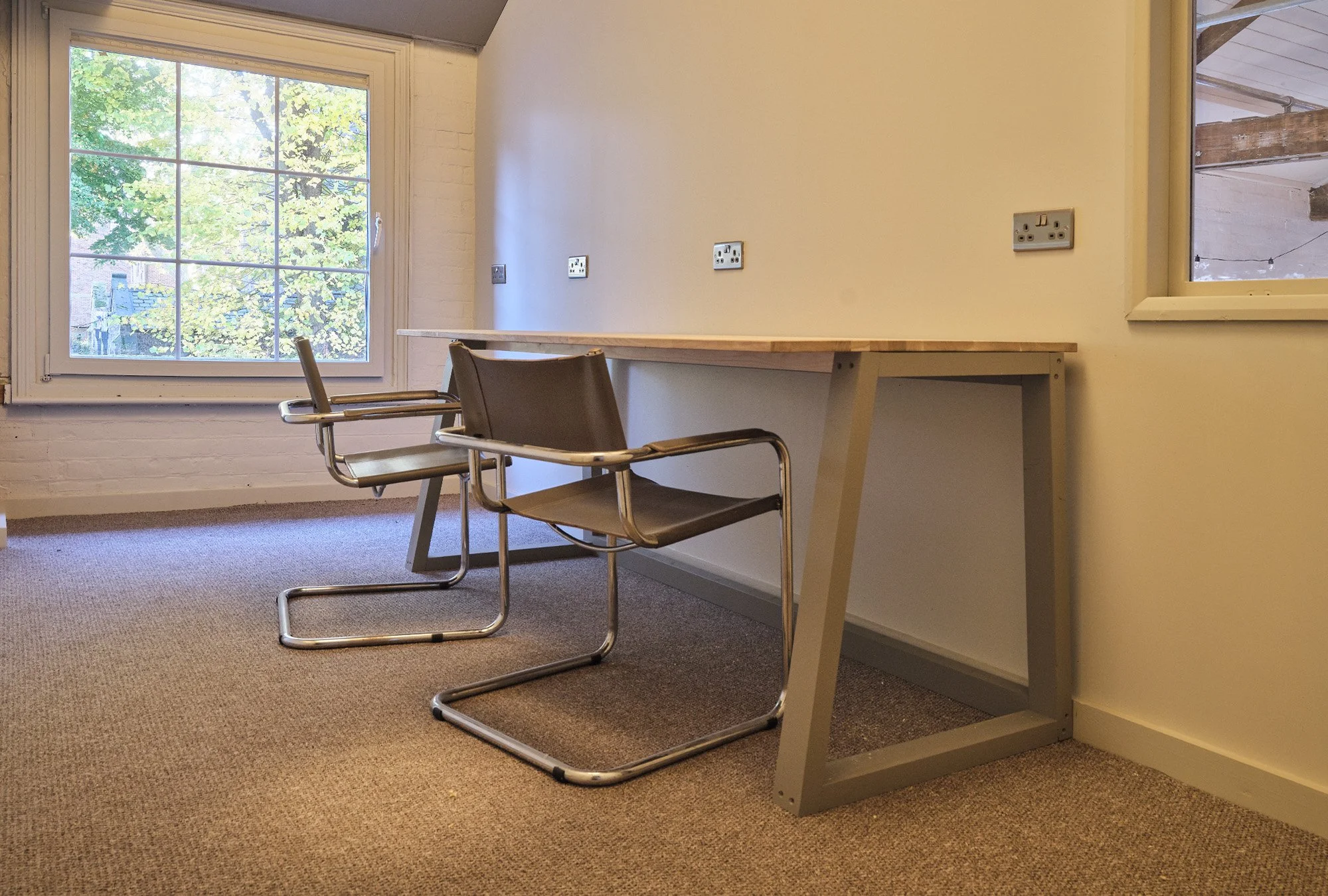 An empty room with a wooden desk, two metal chairs, a window showing trees outside, and multiple electrical outlets on the wall.