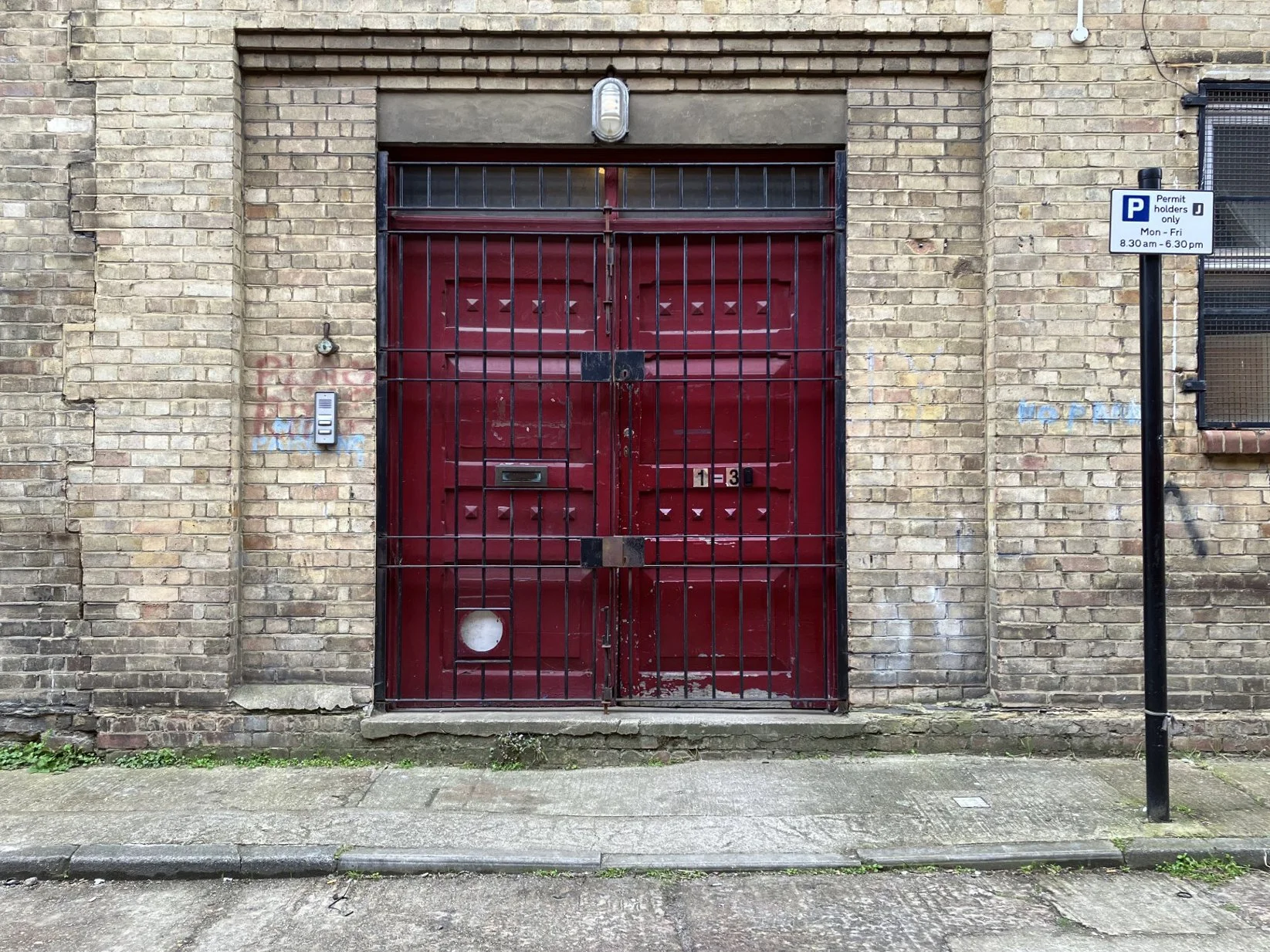 Red metal garage door with barred security gate on brick building, intercom on the left, parking permit sign on the right.