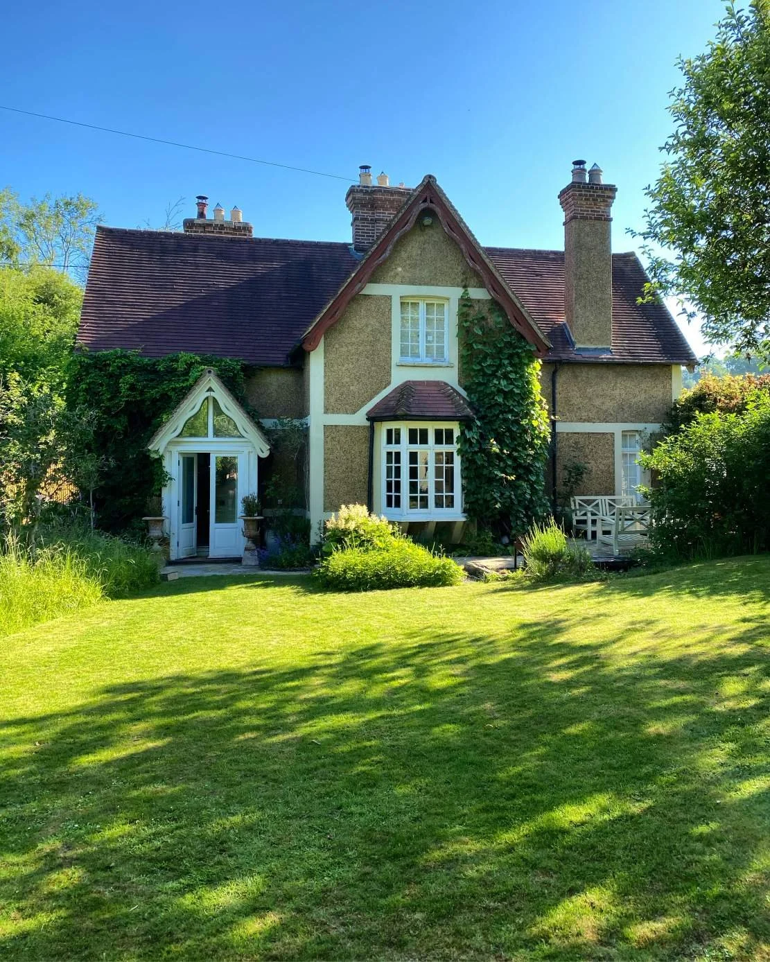 A charming house with a steep roof, featuring a small front porch, bay window, and a large chimney, surrounded by a well-maintained lawn and lush green trees under a bright blue sky.