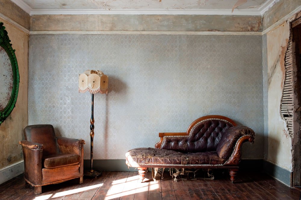An old, worn living room with a brown armchair, a vintage brown leather sofa with torn fabric, a tall floor lamp with a decorative shade, and a damaged wall with peeling paint and exposed wood on one side.