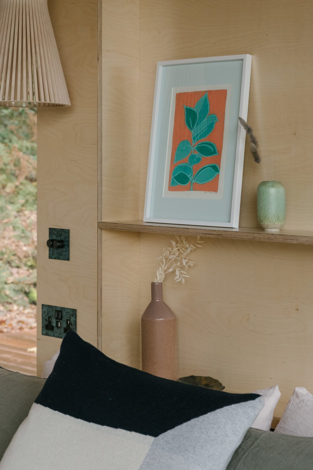 Interior of a room with a beige wooden wall. A framed artwork of a green plant on an orange background is placed on a floating shelf. Next to it, a small green ceramic vase with a feather is visible. Below, a brown vase with dried white foliage stand
