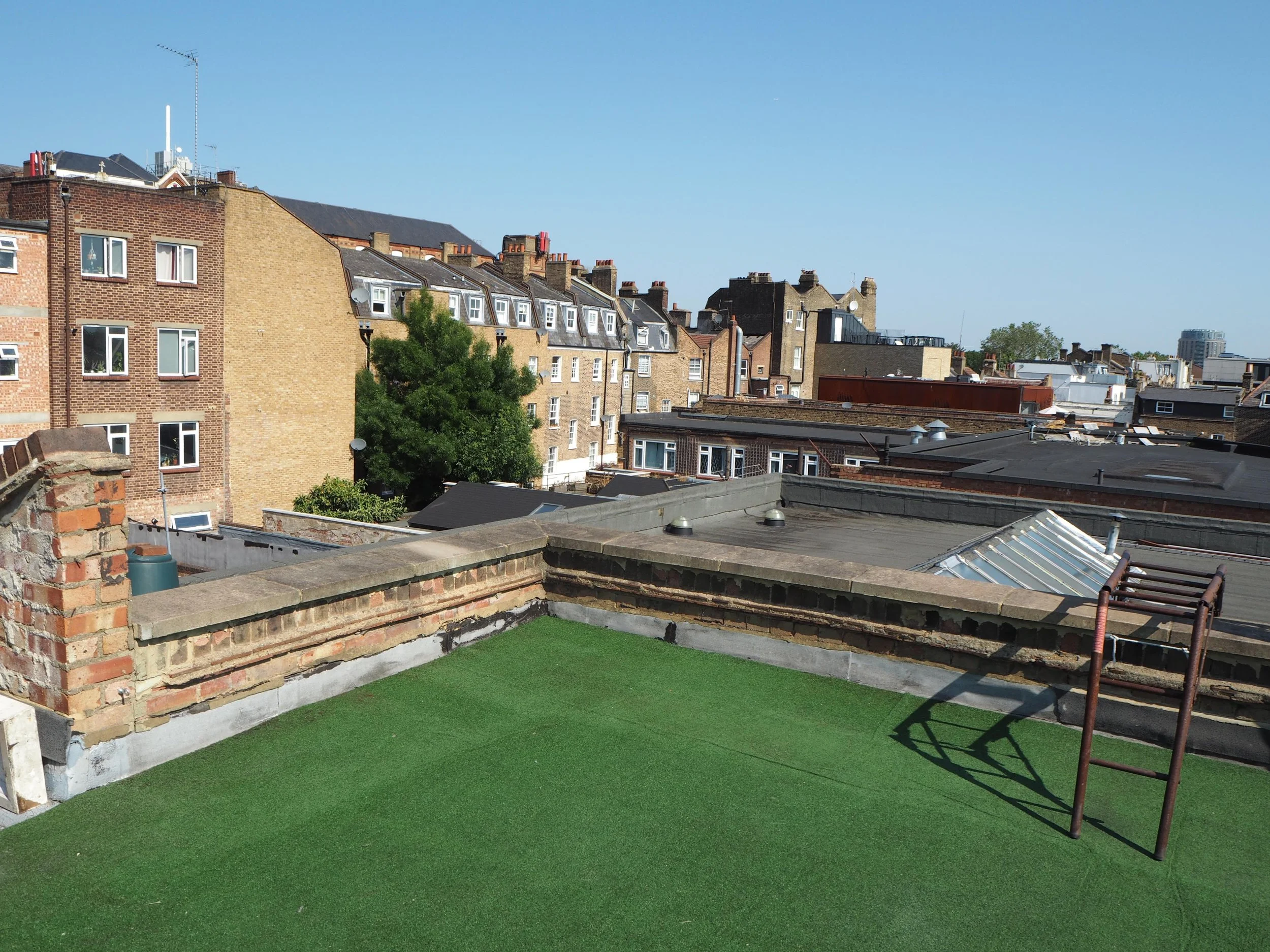 View of city rooftops with a clear blue sky, brick and flat roofs, and some greenery at the edge of the rooftops.