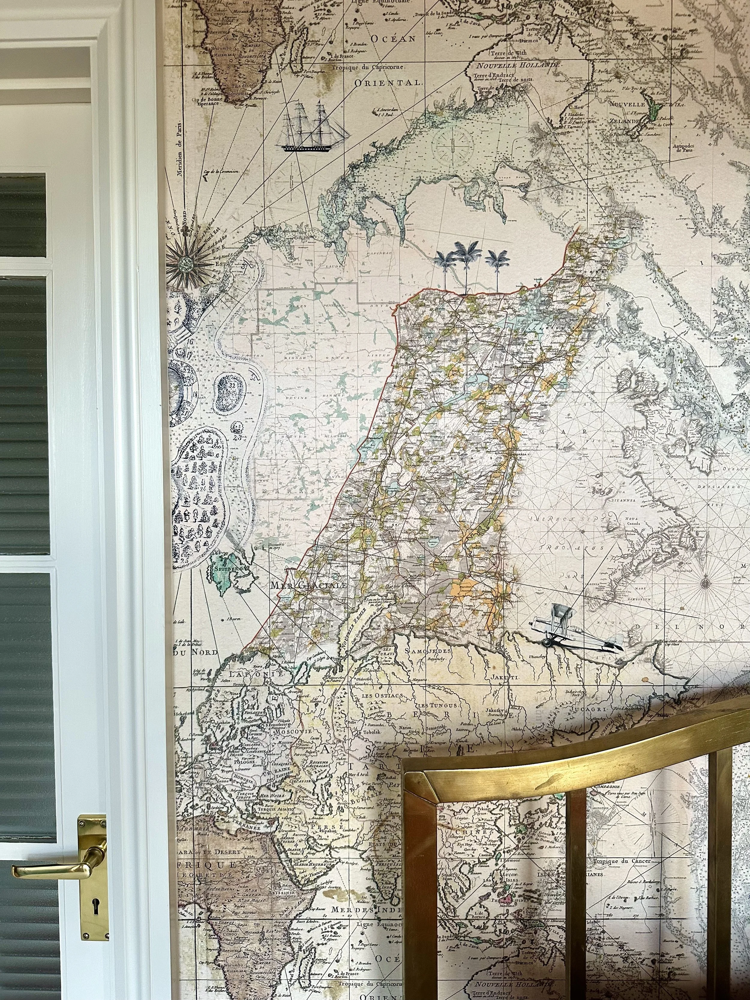 A large vintage-style map of South America and surrounding regions, decorated with illustrations of ships, palm trees, and a compass rose, hanging on a wall next to a white door with a gold handle.