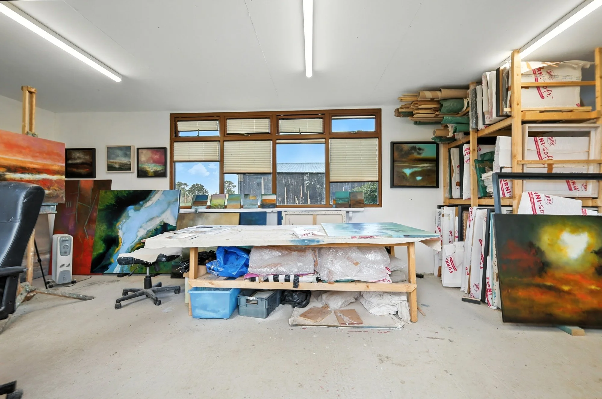 An art studio with large windows, paintings, and art supplies, including canvases, paper, and storage shelves, with a beige floor and white walls.
