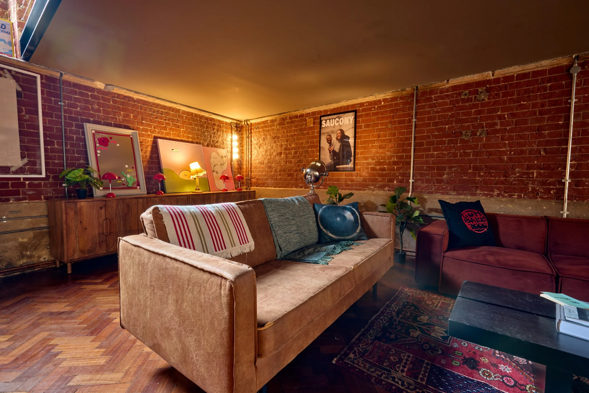 A cozy living room with brick walls, a beige couch with colorful cushions and blankets, a red sofa with a black pillow, a wooden sideboard with lamps and artwork, and a patterned rug on wooden floors.