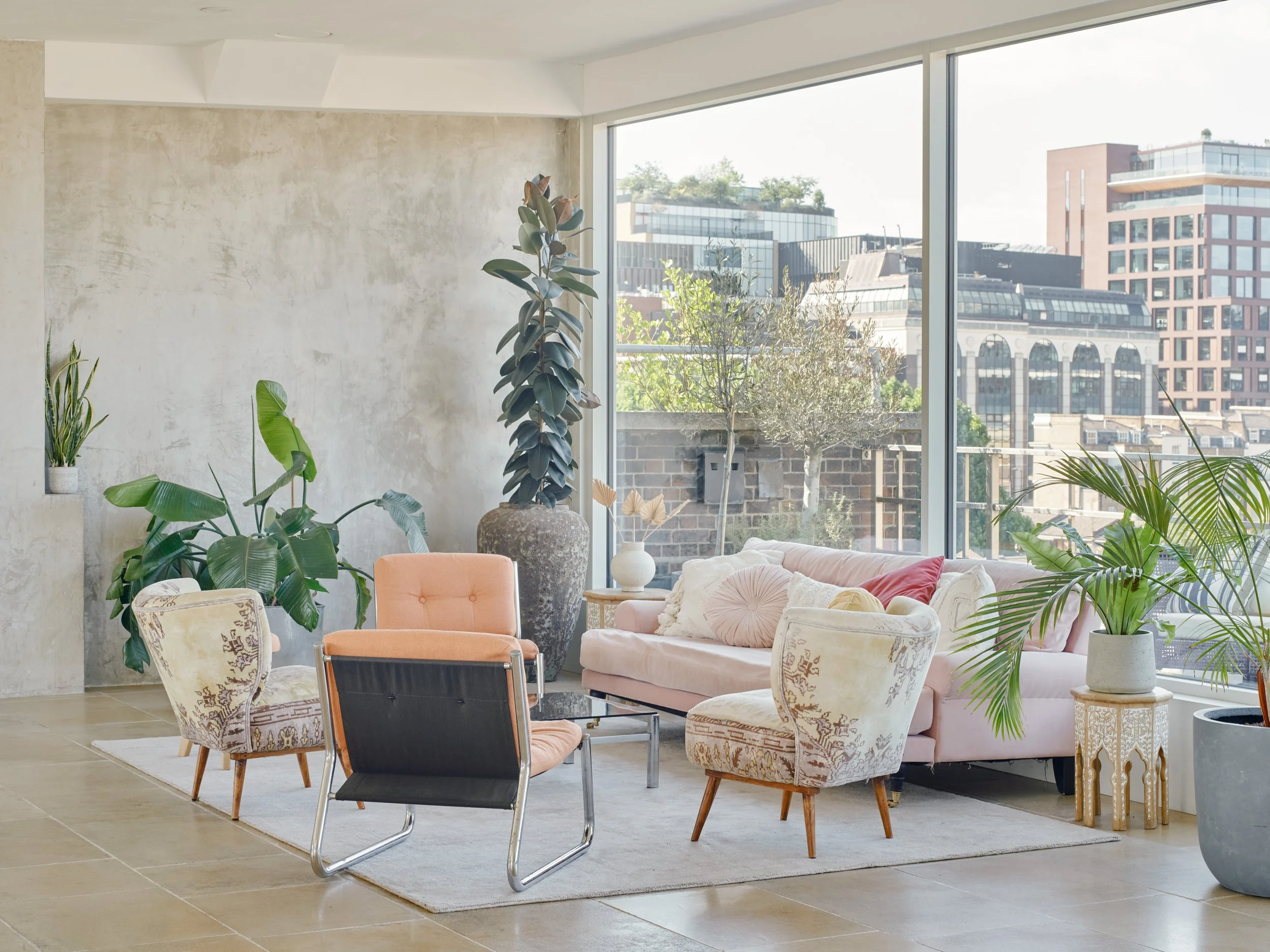 Modern living room with large floor-to-ceiling windows, pastel pink and floral-patterned chairs, and various green indoor plants, overlooking city buildings