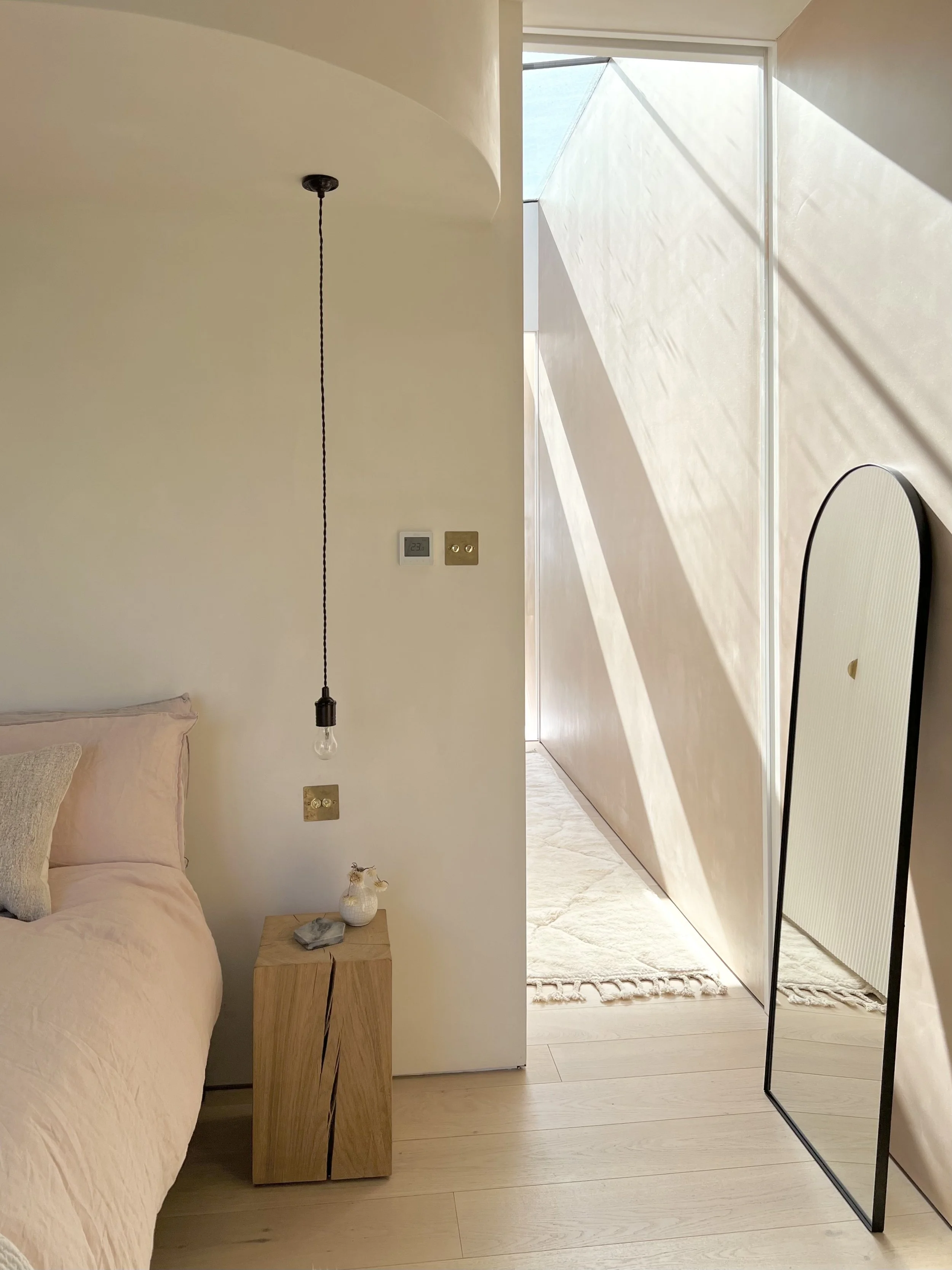 Minimalist bedroom with beige walls, a bed with pale pink linens, a wooden nightstand with a small vase and decorative stones, and a hanging pendant light with an exposed bulb. There are two electrical outlets on the wall, a full-length mirror, and a