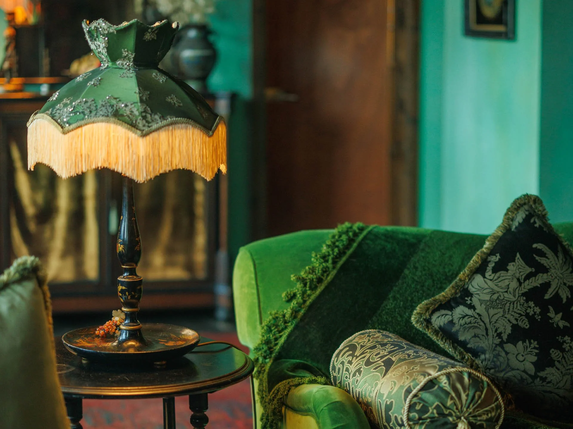 Vintage green velvet sofa with ornate patterned pillows and an antique table lamp with a dark base and a green, fringed lampshade, in a room with green walls and dark wood furniture.