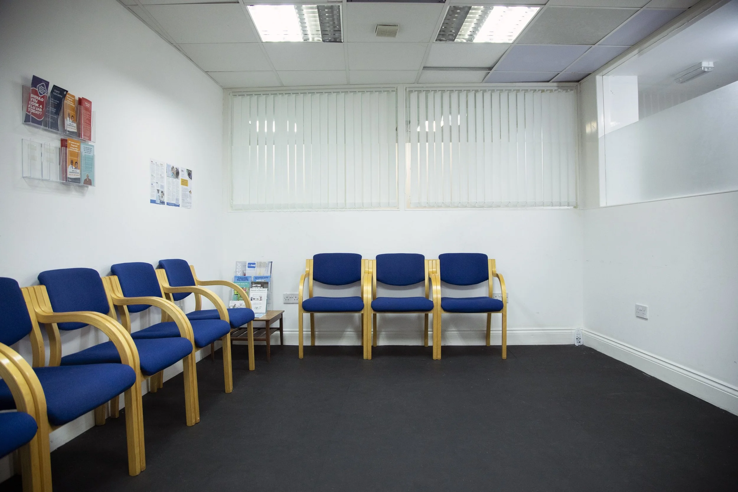 Empty waiting room with blue chairs with wooden armrests, a small table with magazines, white walls, and large window blinds.