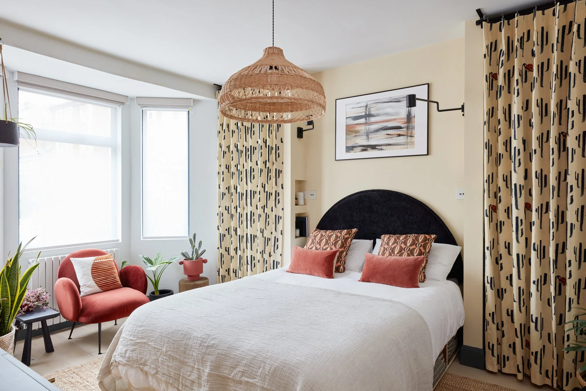 Decorated bedroom with a black headboard, white bed, patterned pillows, and curtains with a cactus design, featuring a woven pendant light and potted plants near a window.
