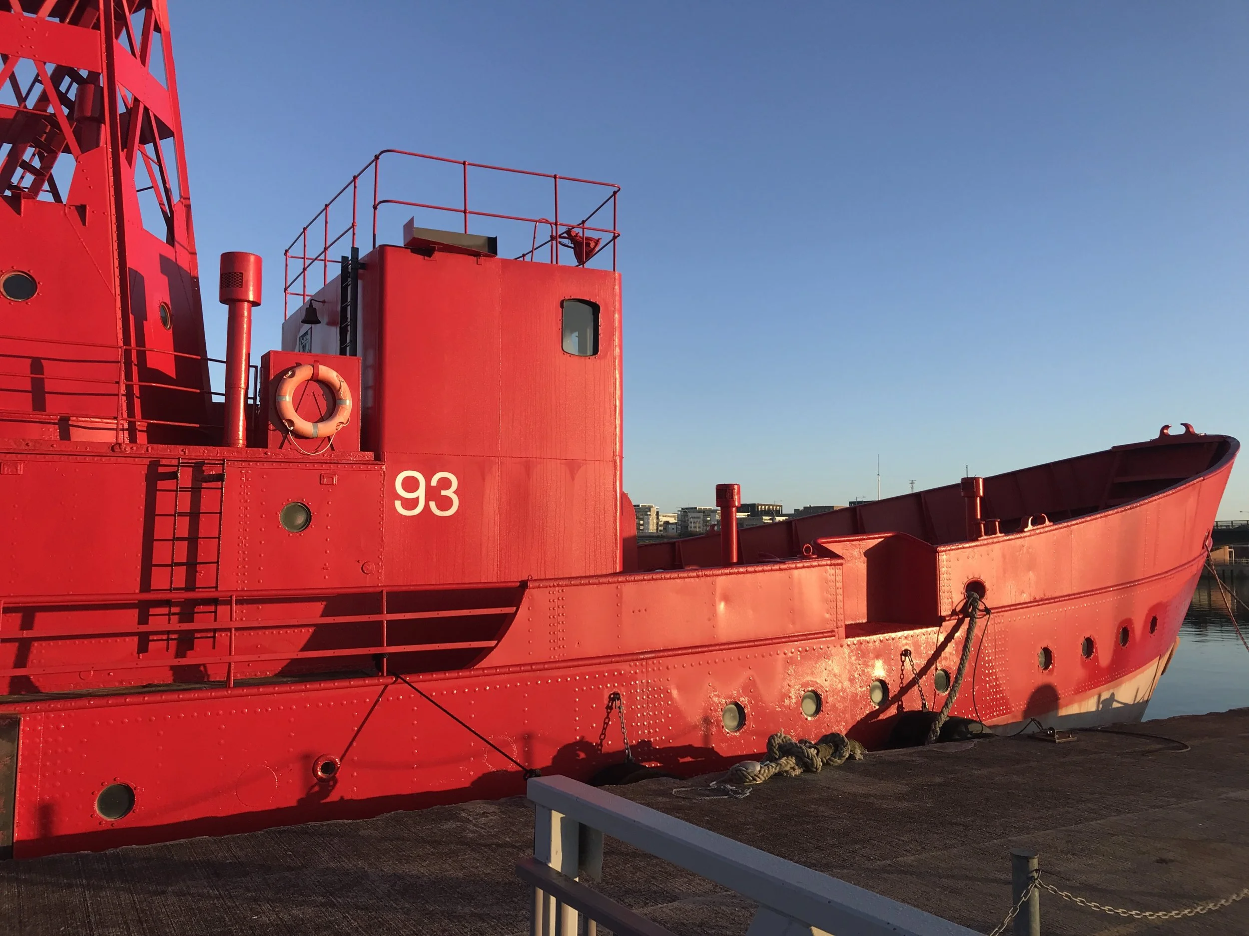 Red ship docked at a harbor with a city skyline in the background during sunset.