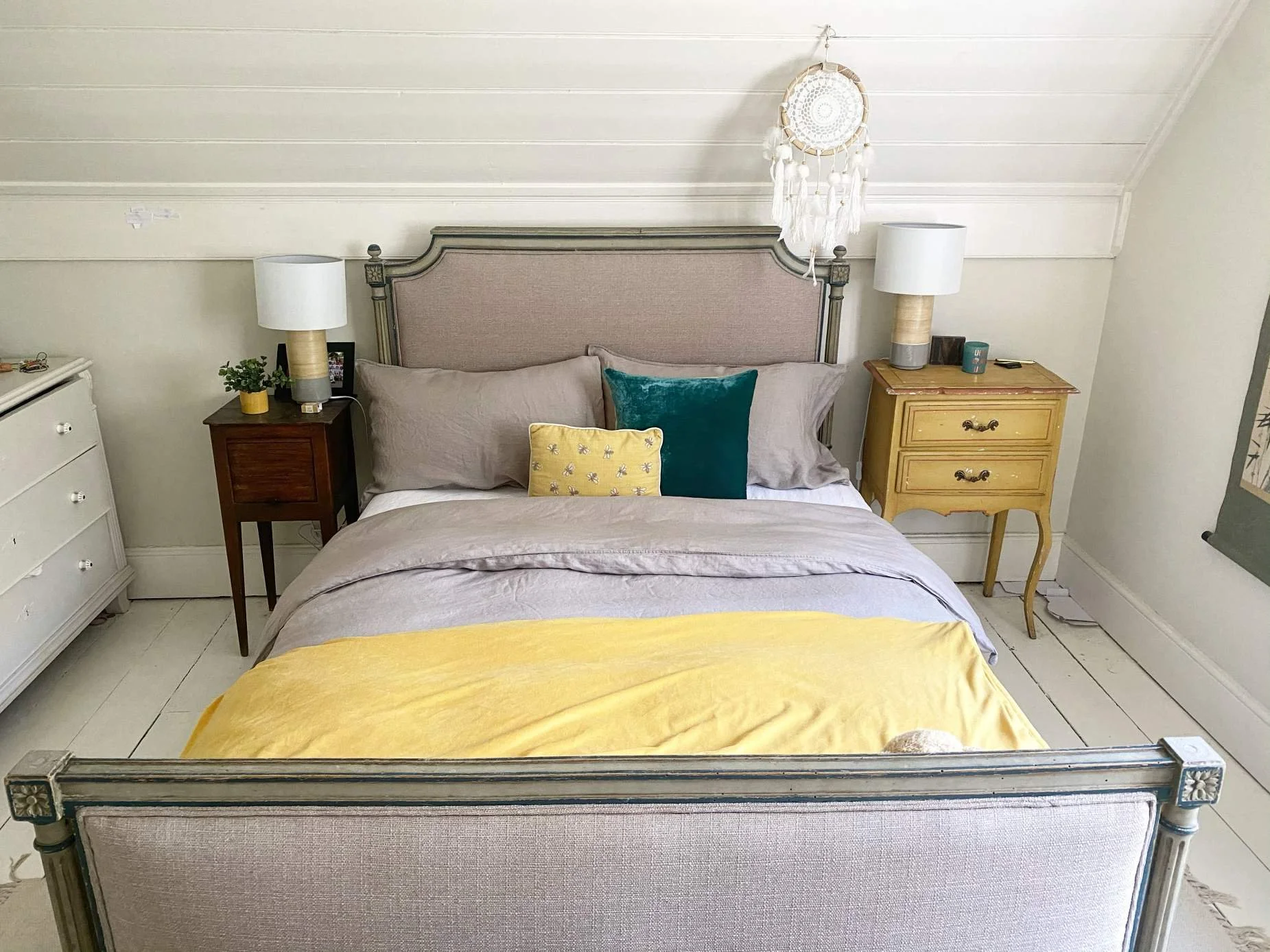 A bedroom with a bed, two nightstands, and a dresser. The bed has a beige headboard, beige sheets, and decorative pillows in teal and yellow. The nightstands are brown and yellow, each with a lamp. A dreamcatcher hangs above the bed on the white wall