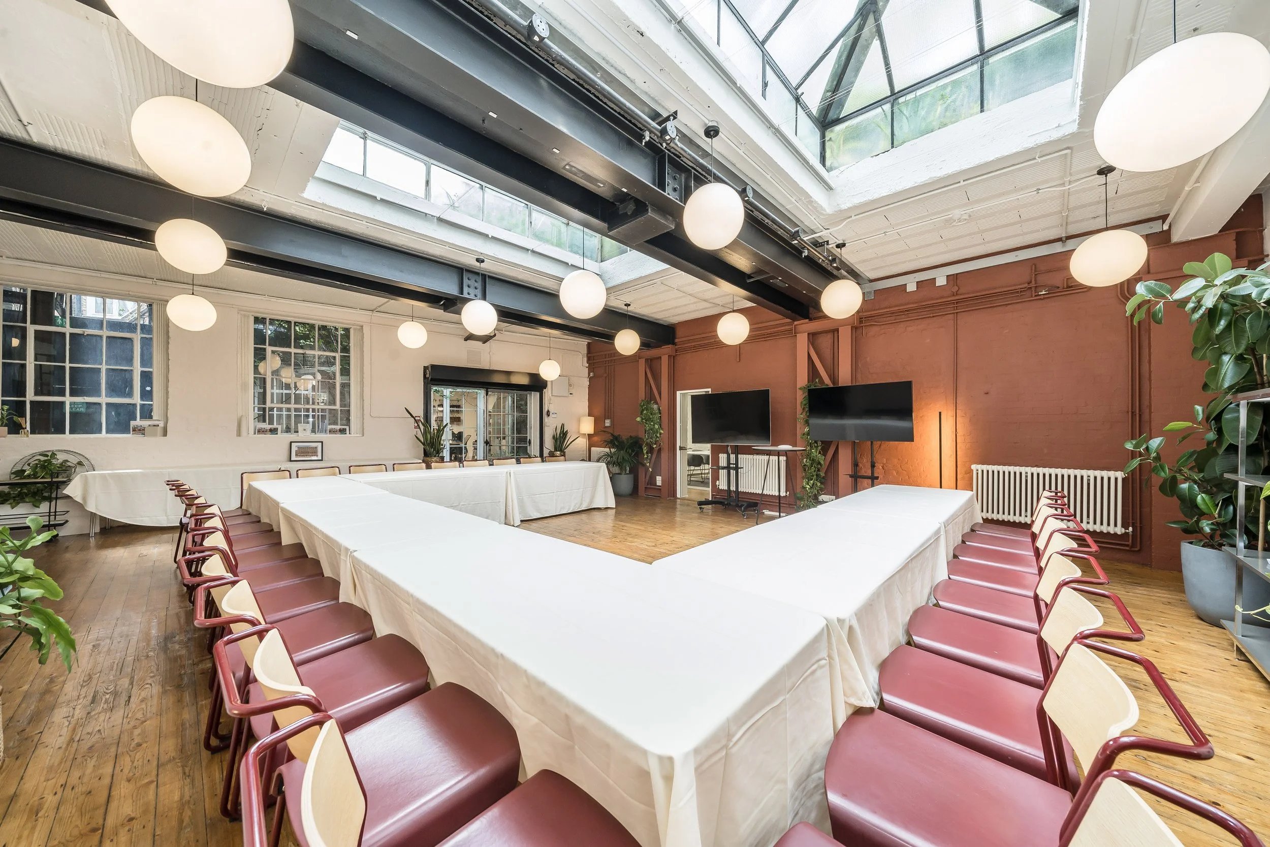 A spacious conference room with a large U-shaped table covered with white tablecloths, surrounded by chairs with maroon seats. The room features large windows, a glass ceiling, and hanging spherical light fixtures. There are plants and two large scre