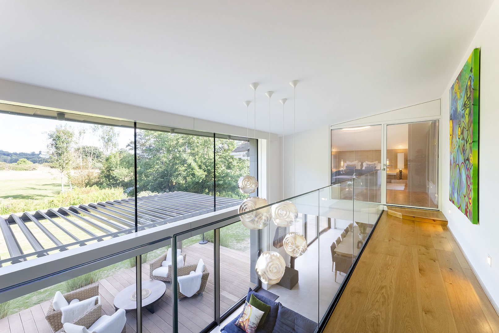 Modern interior house hallway with hardwood floors, glass railing, and large window overlooking outdoor seating and green landscape.