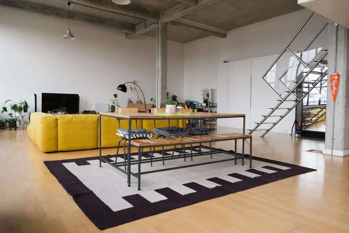 Living room with yellow sofa, black TV, plants, a wooden table with a metal frame, a black and white rug, a white cabinet, and a metal staircase in an industrial-style space with concrete ceiling beams.