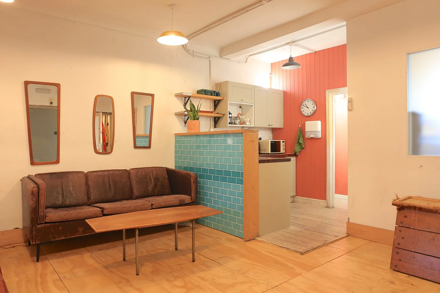 Cozy living room with brown leather sofa, wooden coffee table, three wall mirrors, small kitchen area with teal tiled counter, beige cabinetry, microwave, clock, and dish soap, light wood floor, and warm lighting.