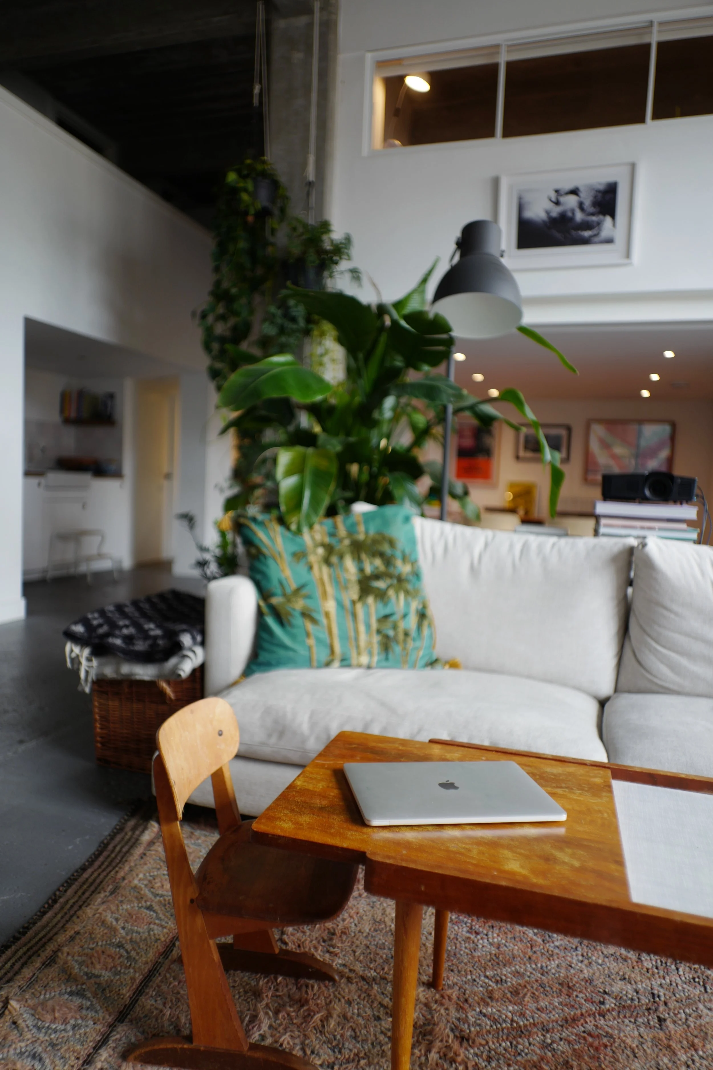 Interior of a modern living room with a white sofa, colorful cushions, a wooden table with a closed laptop, and a chair. There are houseplants, framed artwork, and a pendant light.