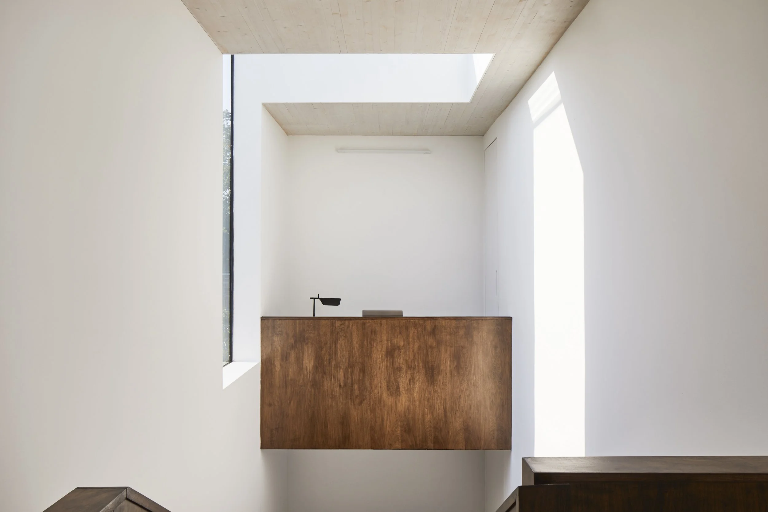 Interior view of a minimalist room with high white walls, a wooden reception desk, a small black desk lamp, and a natural light skylight and window.