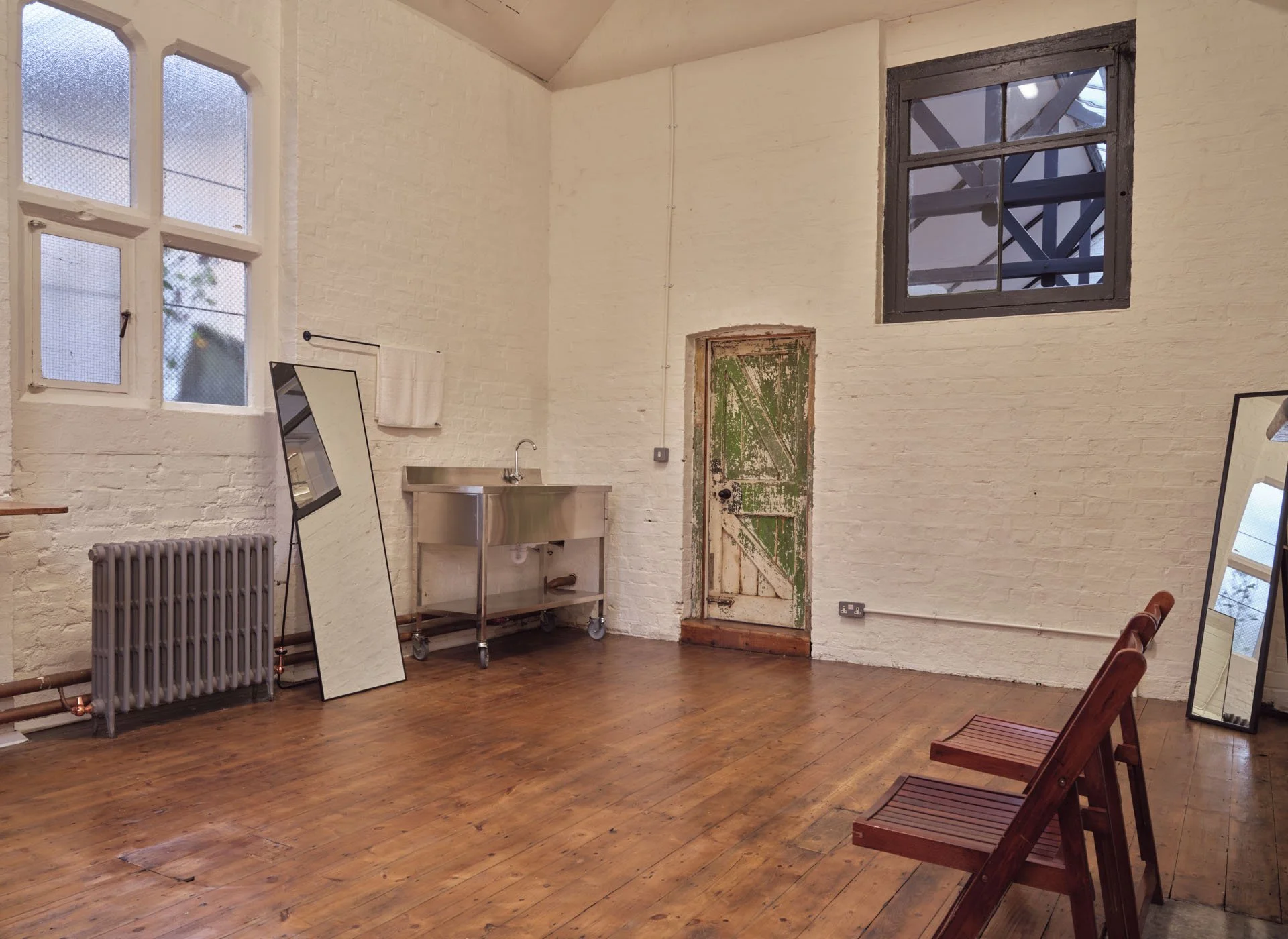 Empty room with white brick walls, wooden floor, two large windows, a mirror, a radiator, a stainless steel sink, two wooden chairs, and an old rustic door.