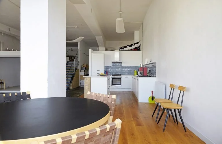 Modern kitchen and dining area with black round table, six chairs, white cabinets, gray backsplash, and wooden flooring.