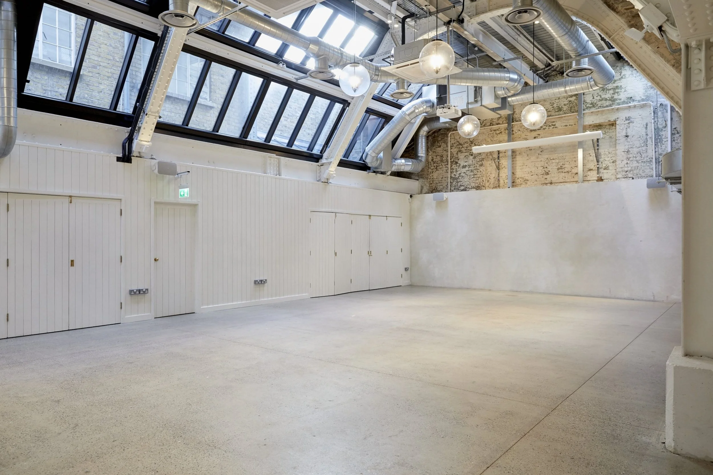 Empty room with high ceiling, large skylight windows, exposed pipes, spotlights, and white paneled walls.