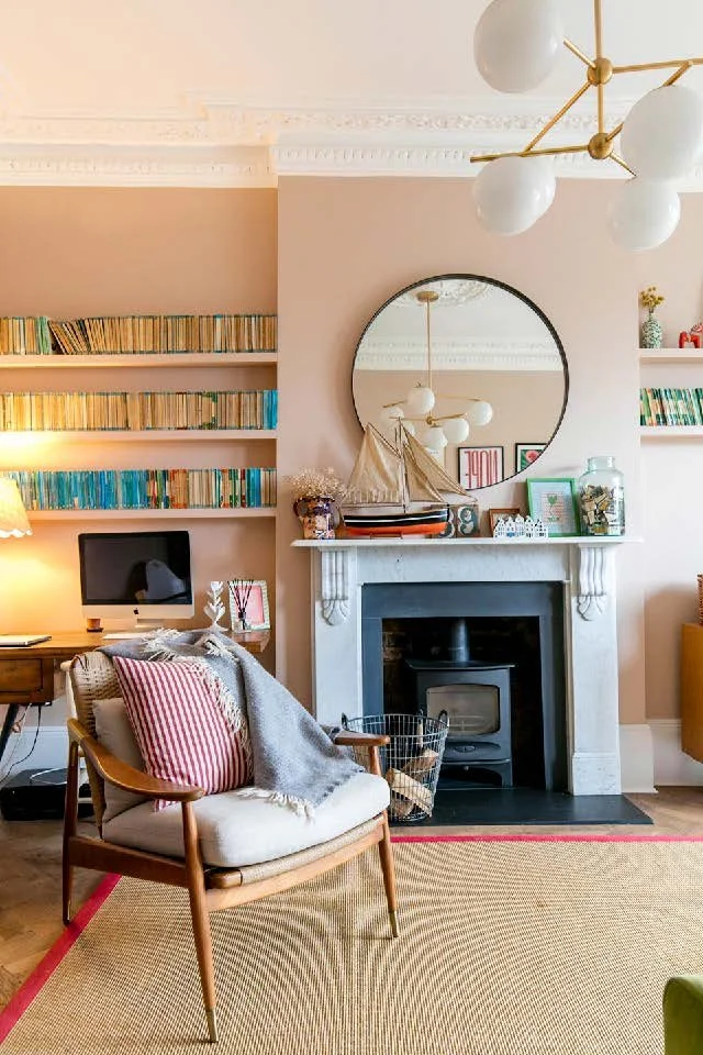 Cozy living room corner with armchair, striped pillow, gray throw blanket, fireplace with decorative items on mantel, round mirror, built-in bookshelves filled with colorful books, and modern chandelier.
