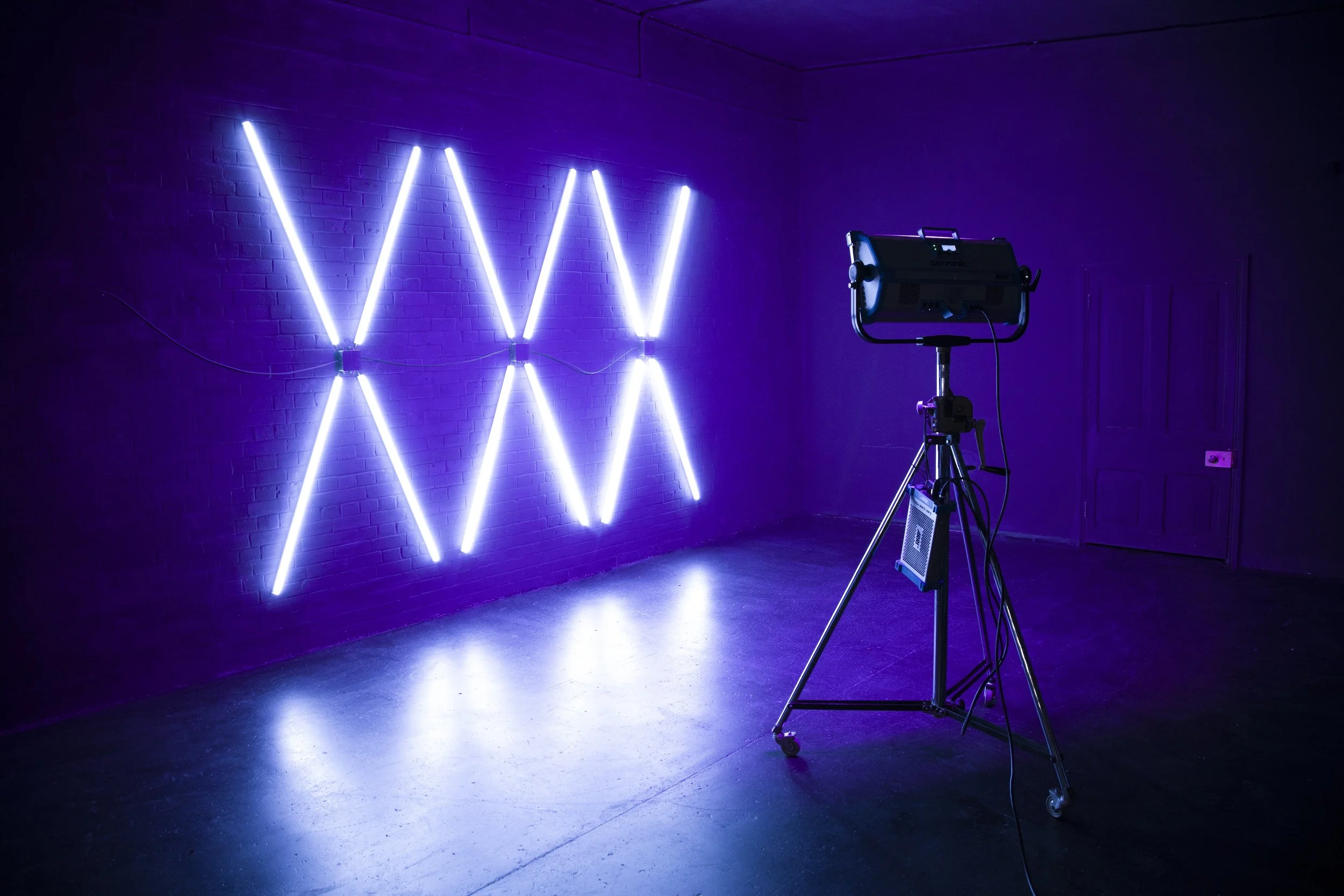 A dark room with purple walls and floor, featuring an art installation of glowing purple and blue neon lights arranged in the shape of three V's and three W's on the wall. A professional light on a stand is aimed at the wall.