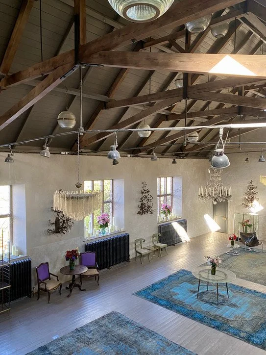 Spacious room with high wooden ceiling, chandeliers, and large windows with flower arrangements, some chairs, and a glass table on a patterned rug.