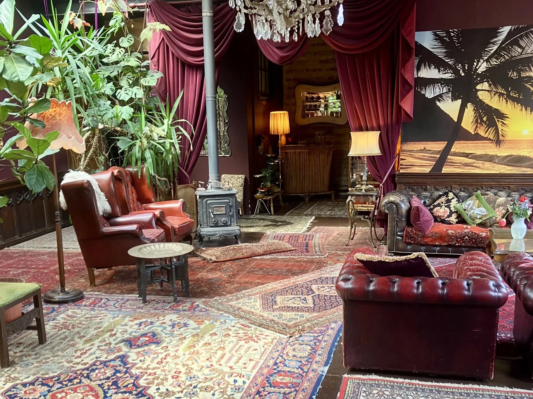 A cozy, vintage-style living room with antique leather sofas, Persian rugs, plants, and a mural of a tropical sunset with palm trees.