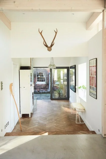 Interior of a room with a deer head wall mount, a white cabinet, a broom, a lit chandelier, a small bench with a pillow, and a poster on the wall. Large windows and a door leading outside are visible.