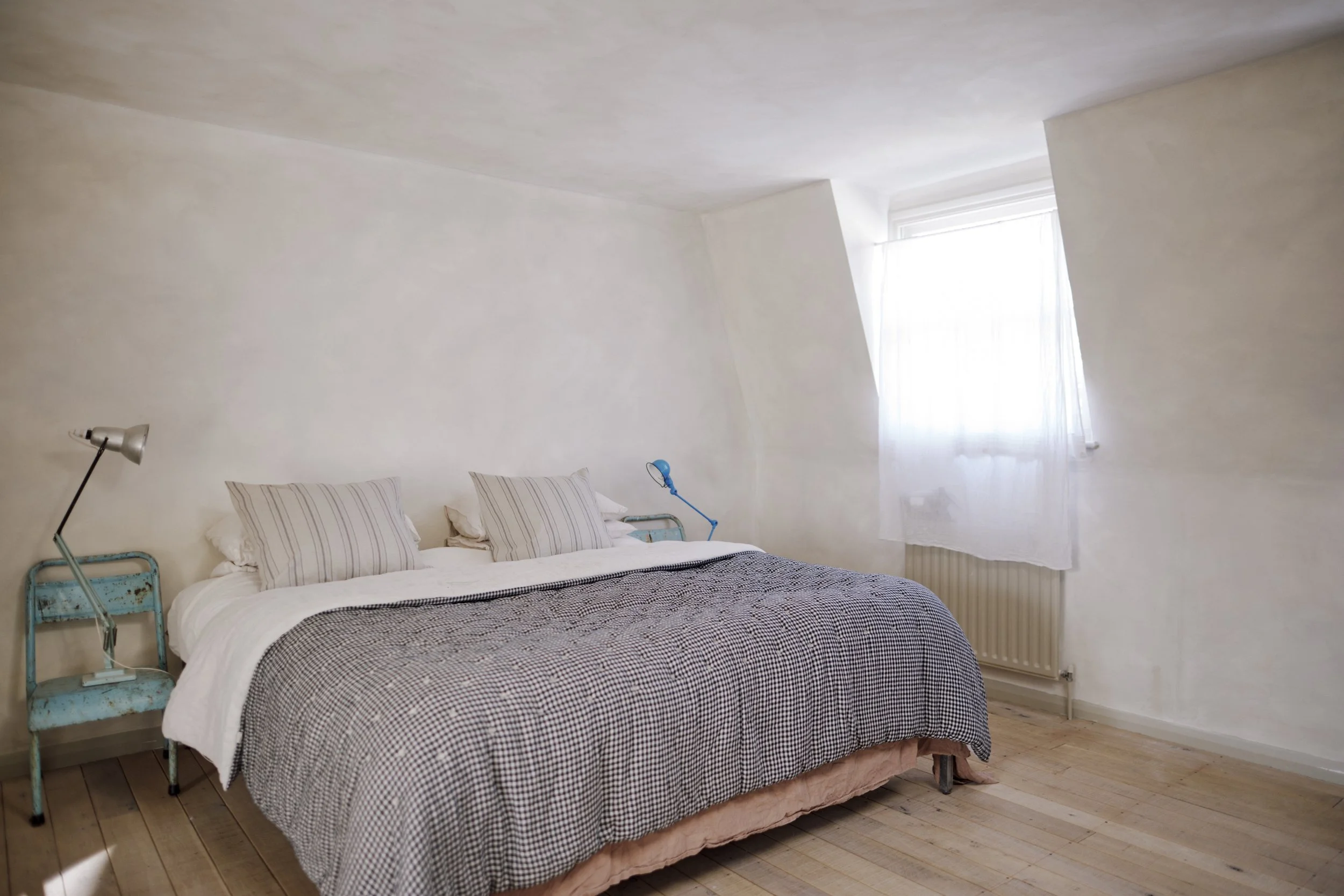 A minimalist bedroom with a large bed, featuring striped pillows and a checkered blanket. Two vintage blue lamps are placed on either side of the bed. A window with a white curtain lets in natural light, and the room has light-colored wooden floors a