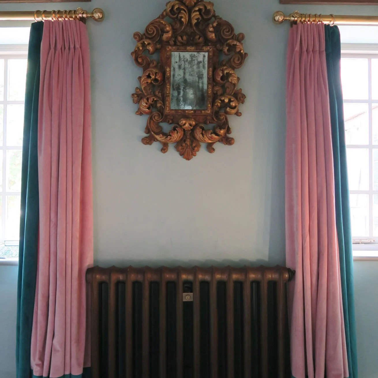 Decorative mirror with an ornate gold frame hanging on a light blue wall between two pink and teal curtains.