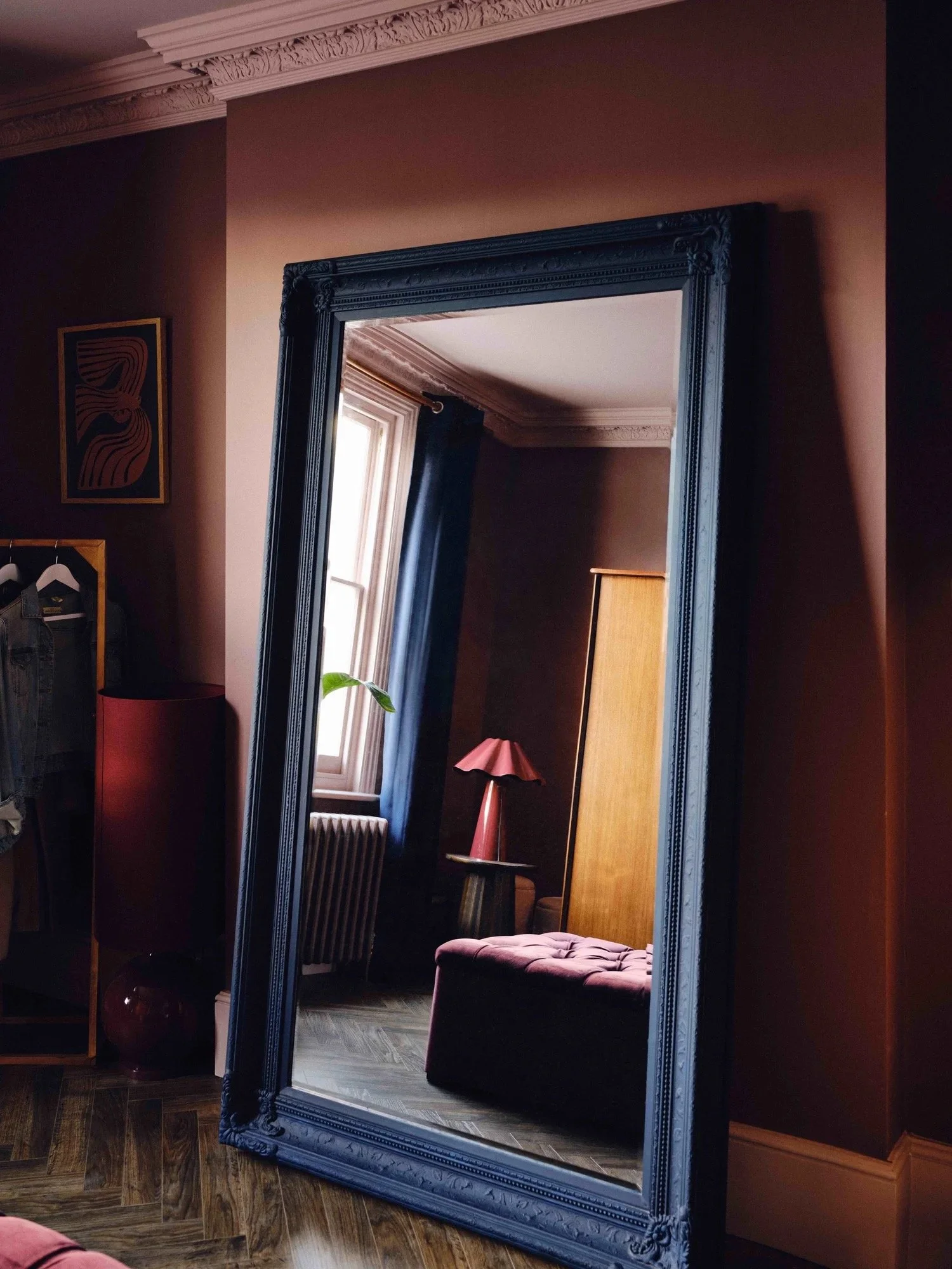 A tall, ornate black-framed mirror reflecting a cozy room with a window, blue curtains, a pink table lamp, a wooden wardrobe, and a pink cushioned chair.