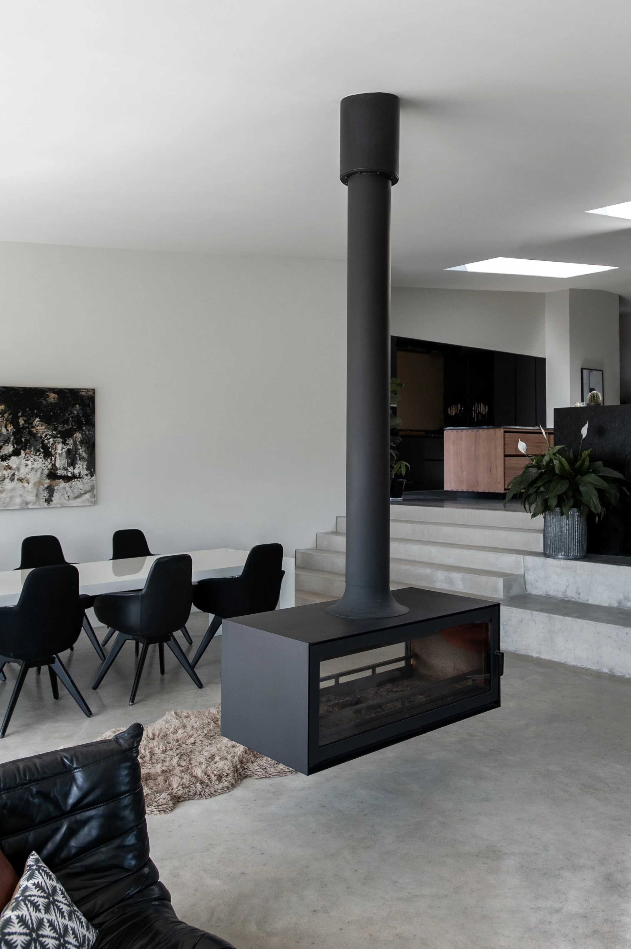 Modern living room with a black suspended fireplace, concrete floors, and minimalist decor, including a dining table with black chairs and a large potted plant.