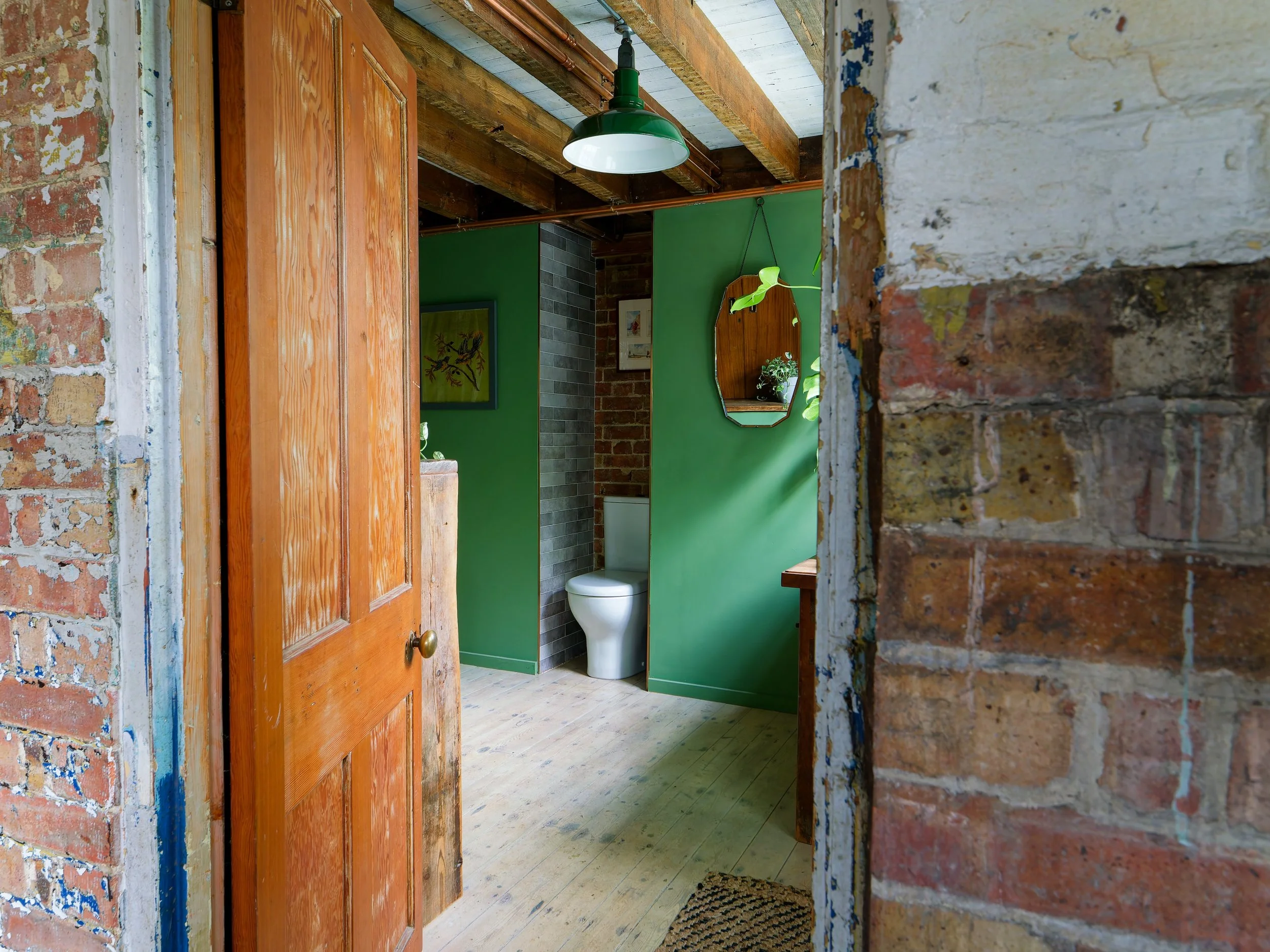 Interior of a bathroom with green walls, visible toilet, mirror, and framed art, with rustic brick and wood accents.