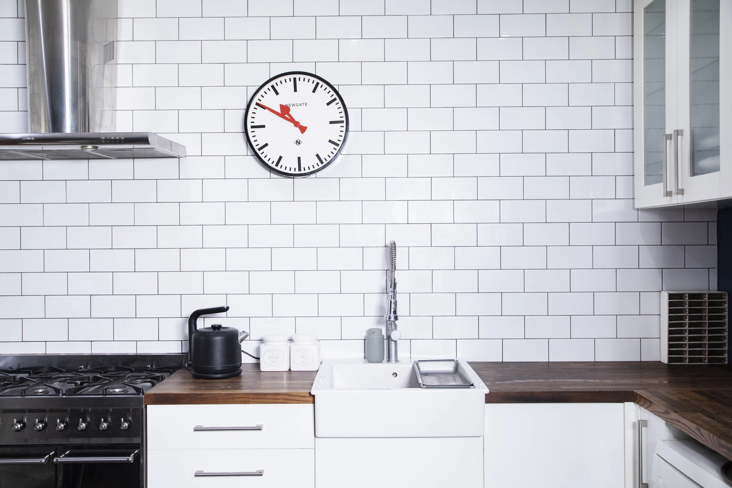 Modern kitchen with white subway tile backsplash, a wall clock, a black kettle, white jars, a dishwasher, and a dark wooden countertop.