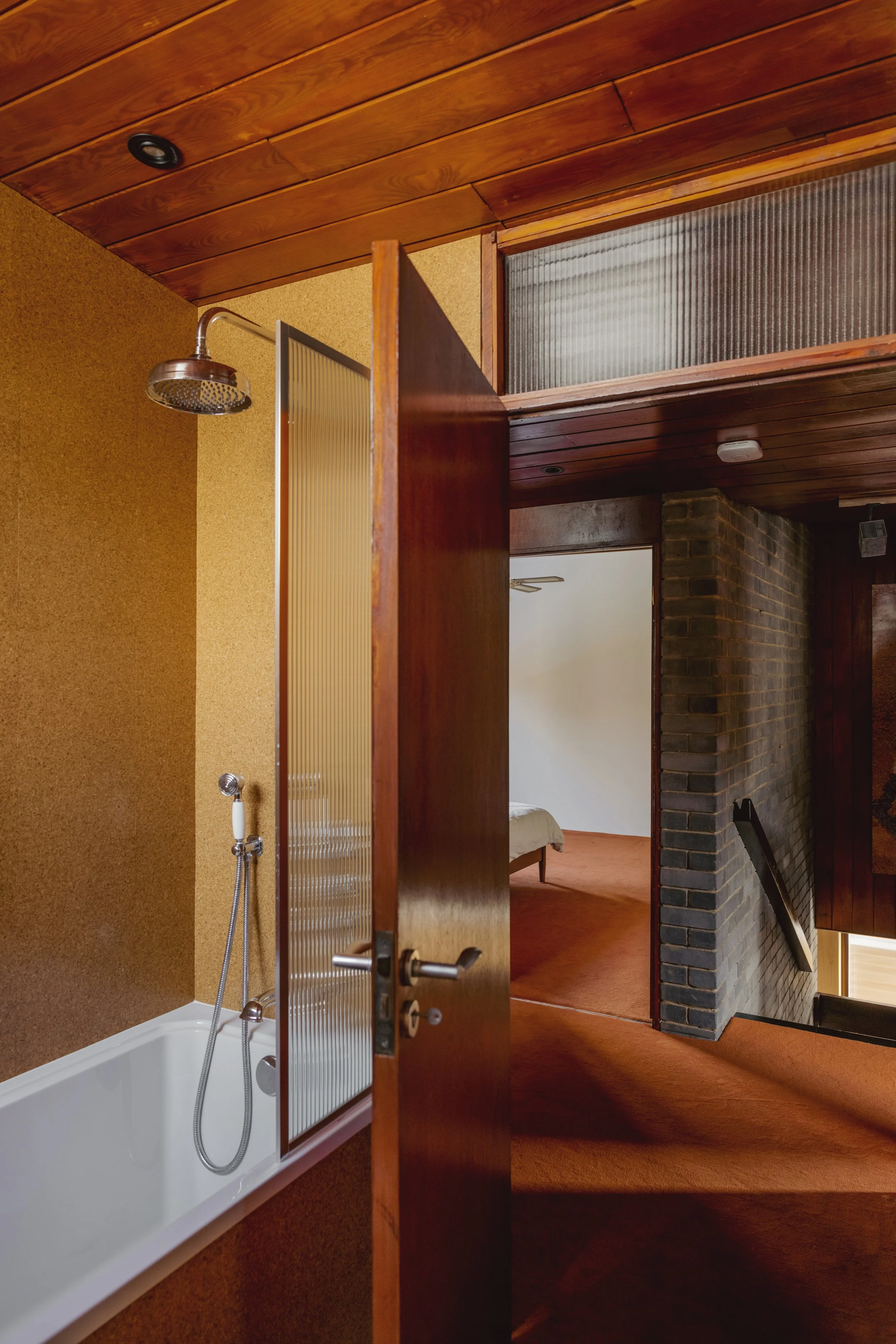 Interior view of a bathroom with a shower, open wooden door, and a bedroom with a bed and brick fireplace in the background.