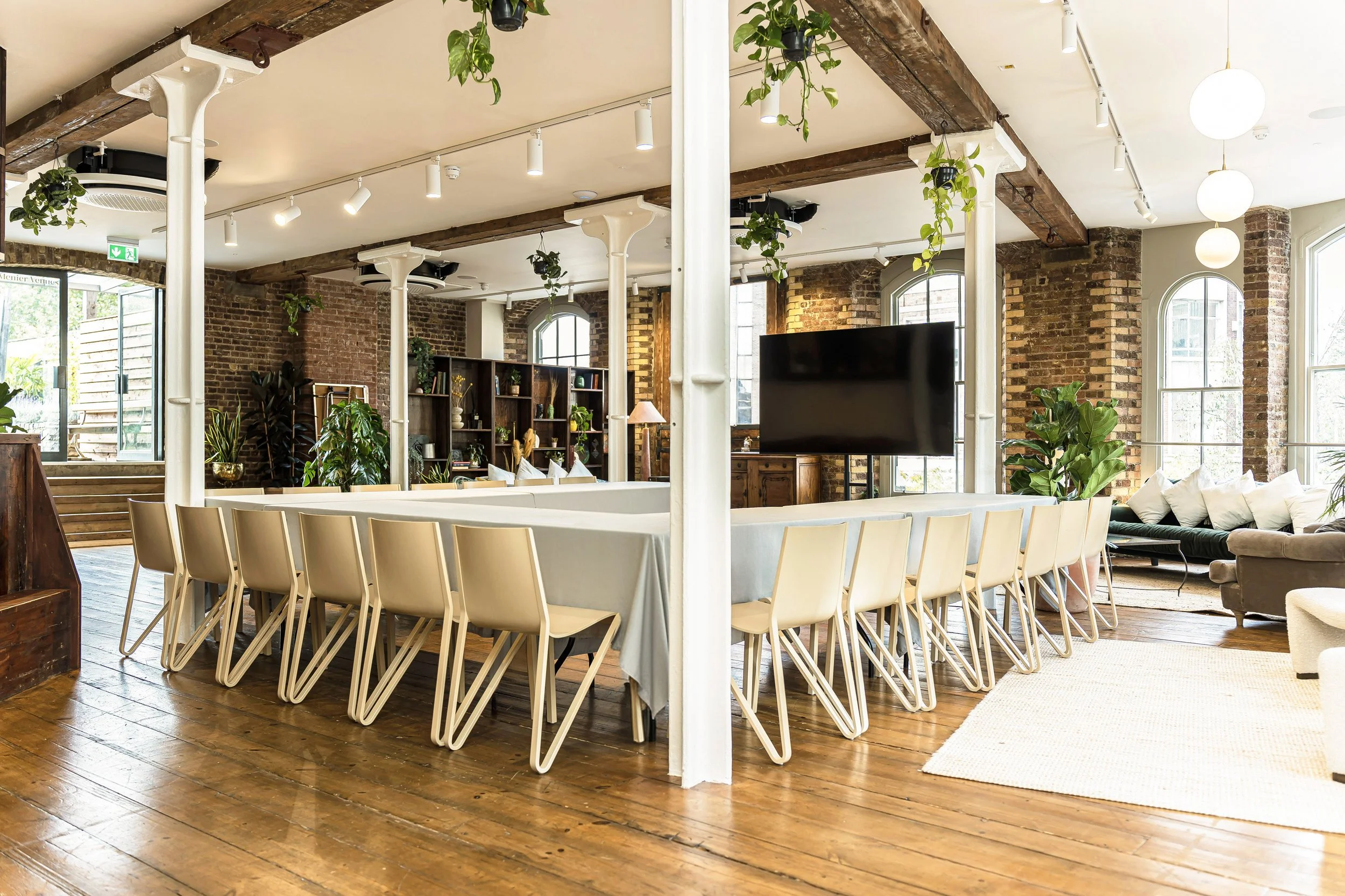 A spacious, industrial-style interior with brick walls, wooden beams, and large windows. There is a long, rectangular table with chairs, a large TV, potted plants, and seating area with sofas and cushions.
