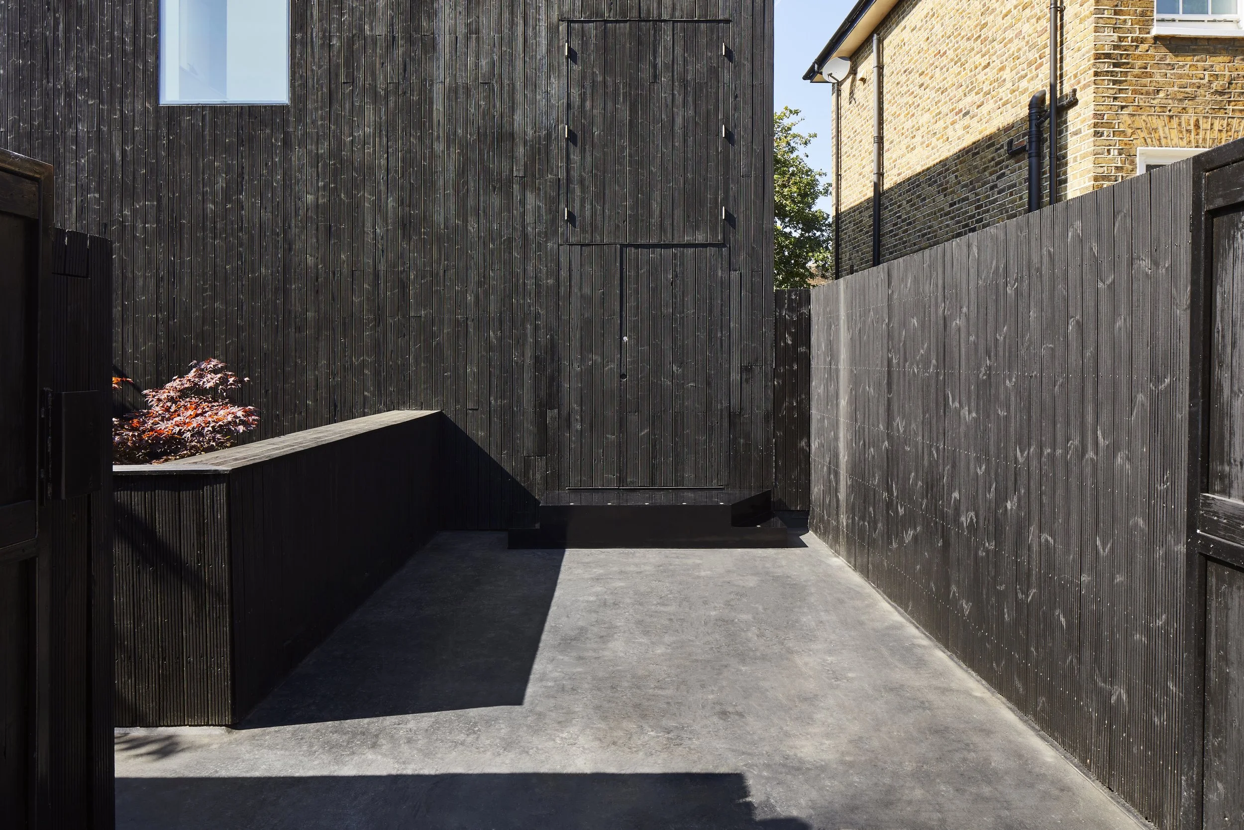 A small outdoor patio area enclosed by black wooden fences with a dark building wall on one side and a brick house wall on the other. There is a concrete floor and a raised garden bed with a small red-leafed plant on the left side.
