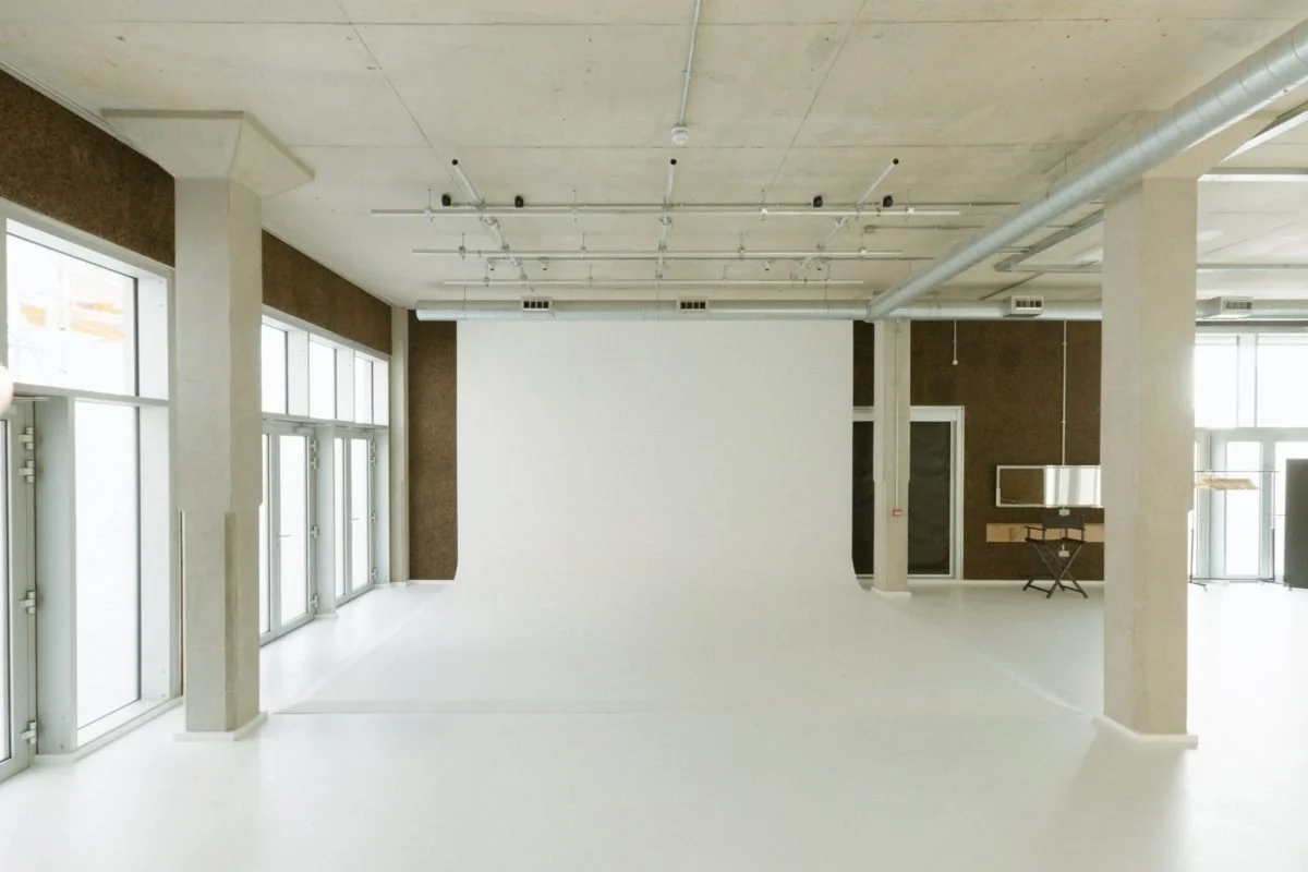 Empty photography studio with a white backdrop, large windows, and industrial ceiling vents.